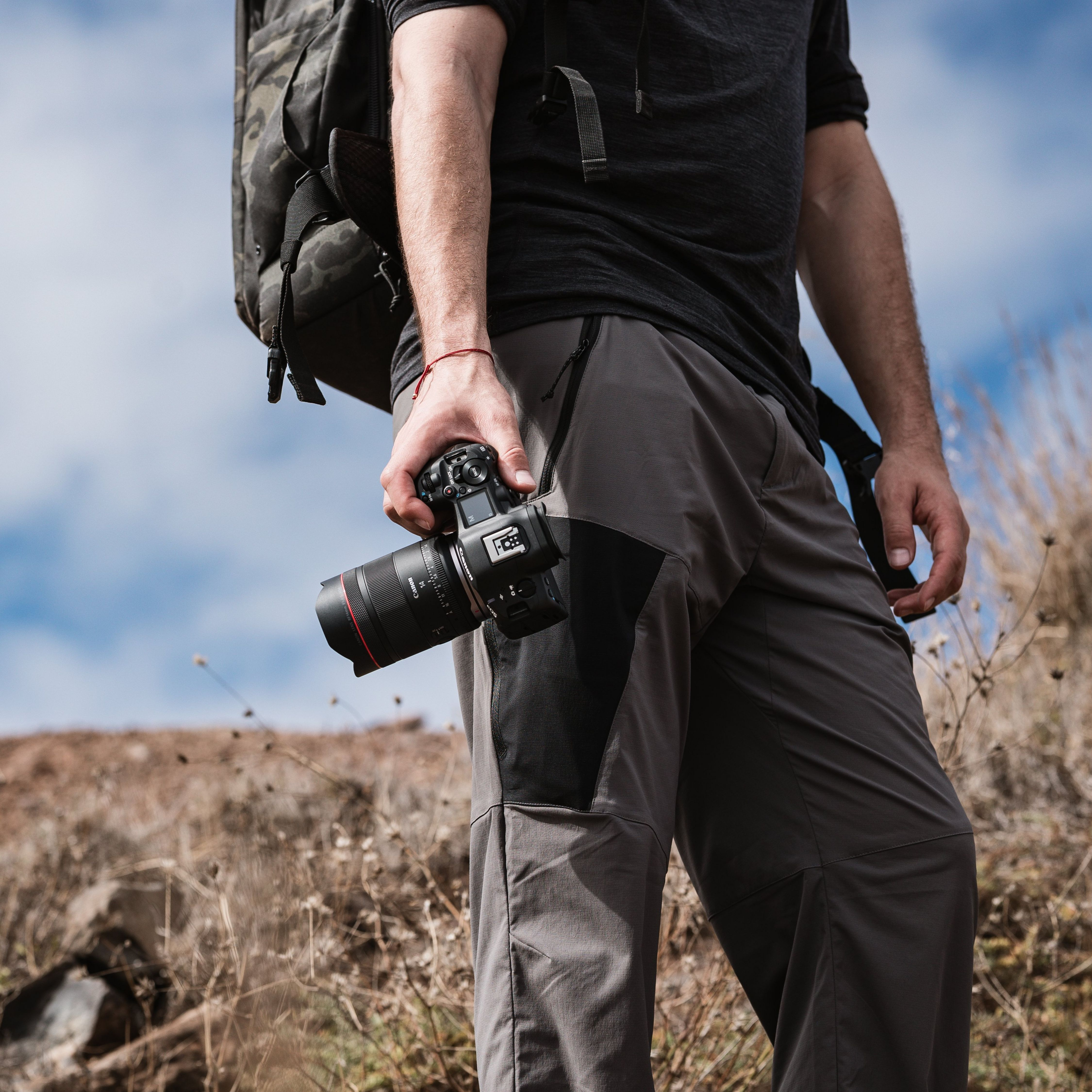 Un homme avec un sac à dos tient un appareil photo, portant un pantalon de randonnée.