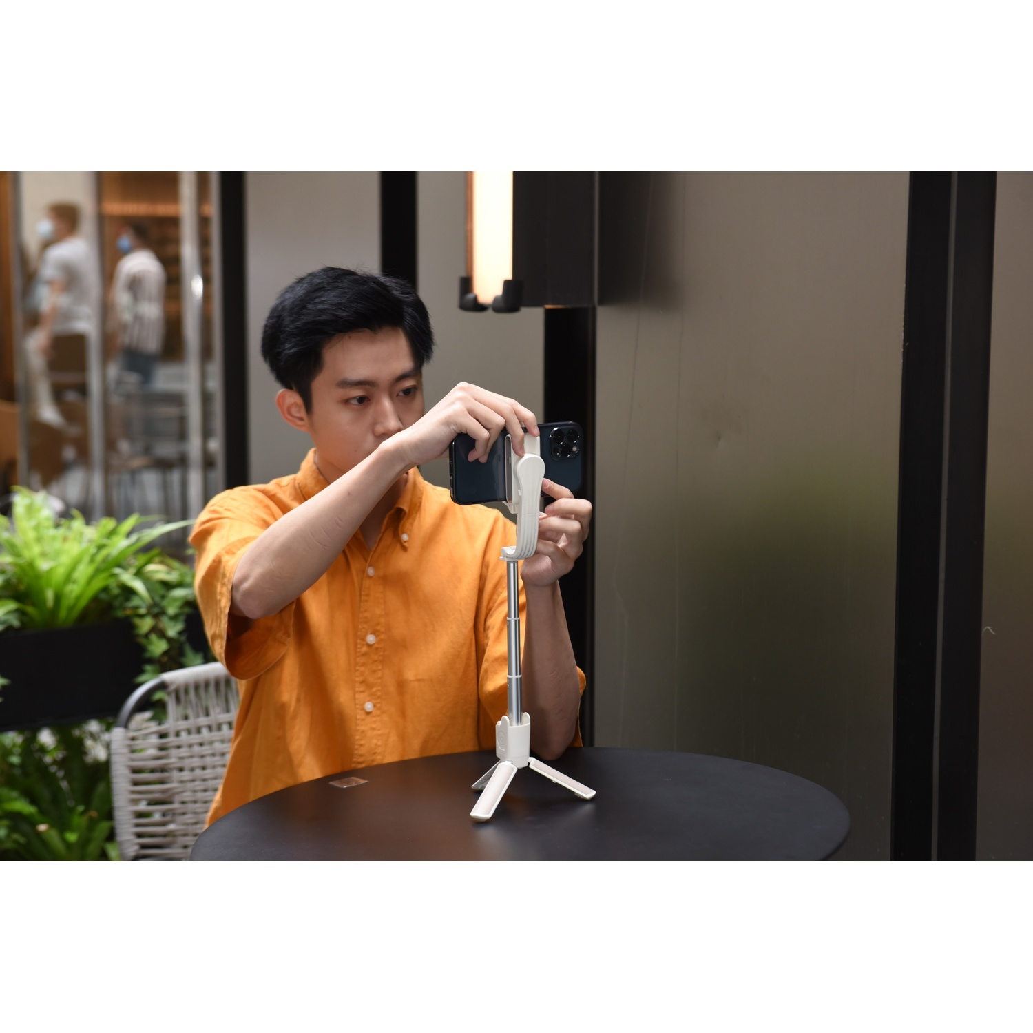 Uomo in camicia arancione tiene uno smartphone su un treppiede gimbal bianco a un tavolo.