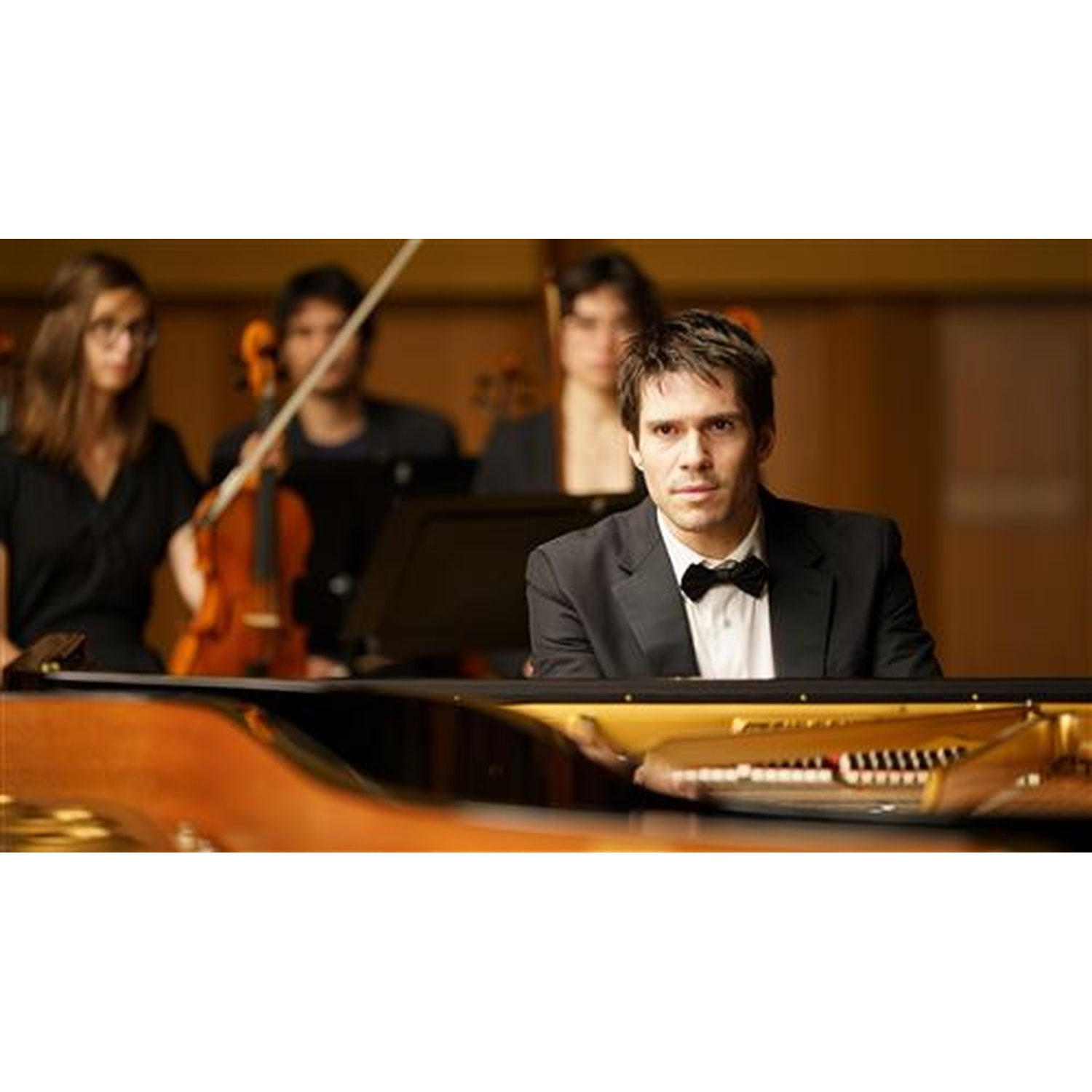 Un homme en costume joue du piano pendant qu'un orchestre se produit en arrière-plan.
