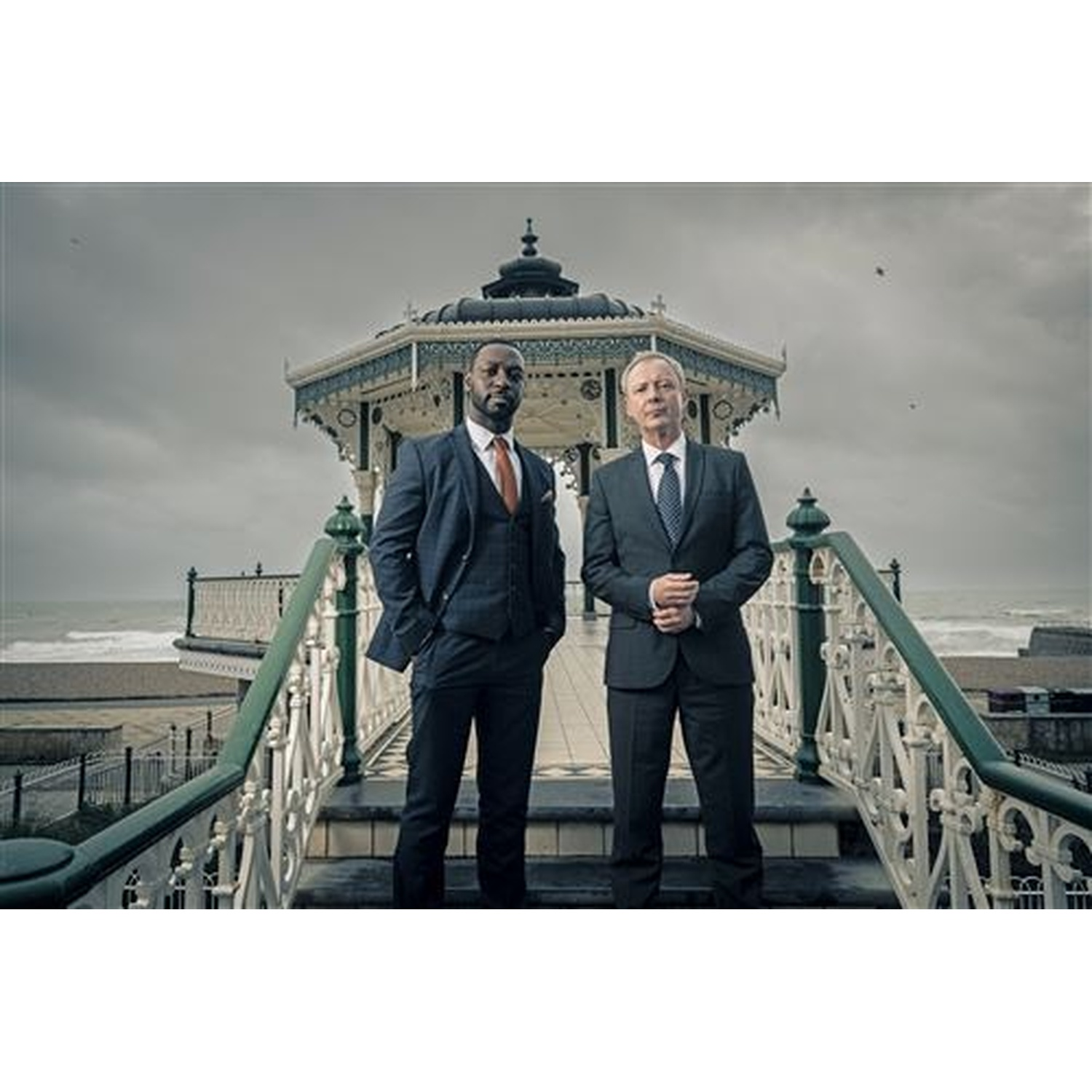 Two men in suits stand on stairs before a seaside bandstand.