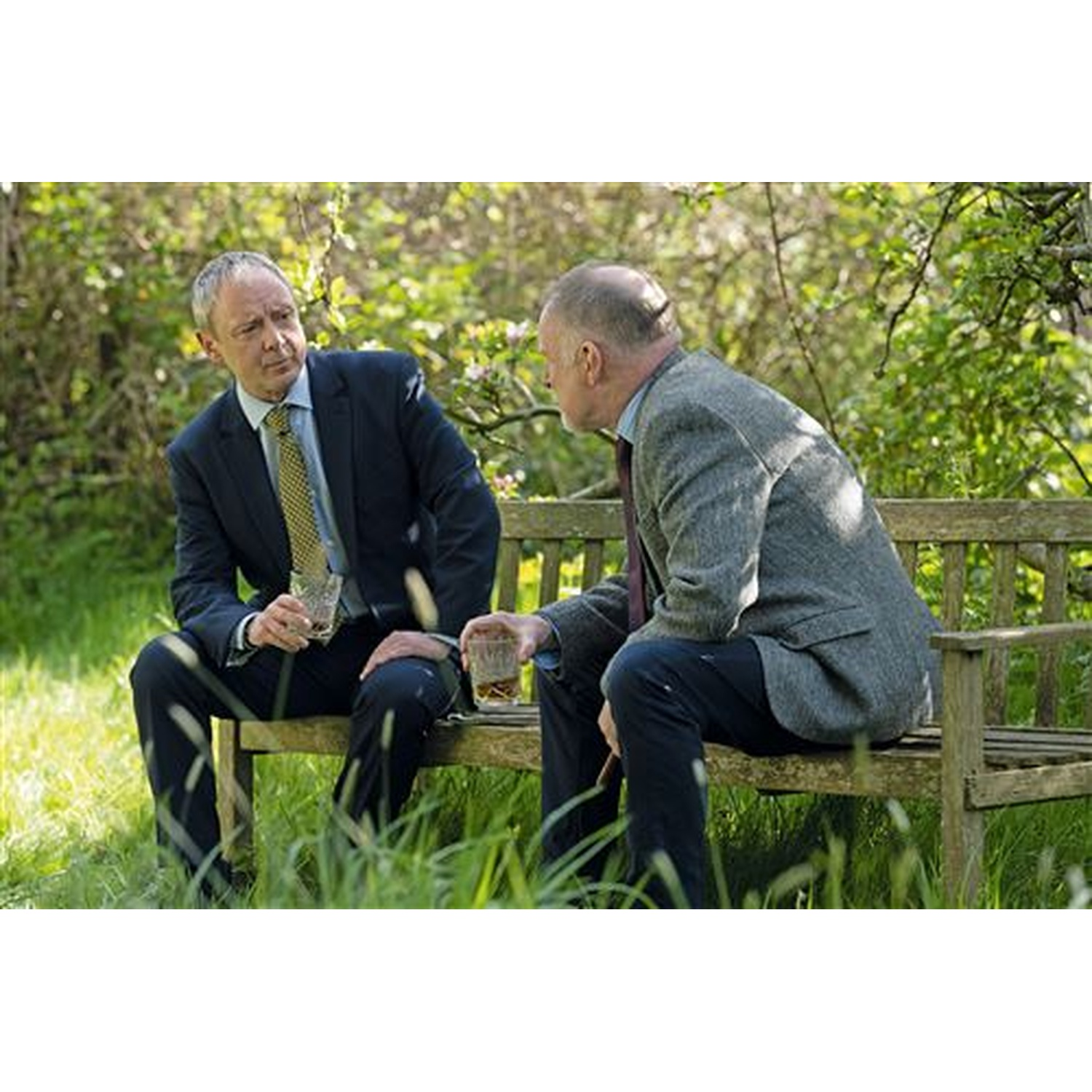 Two men sit on a wooden bench outdoors, holding glasses.