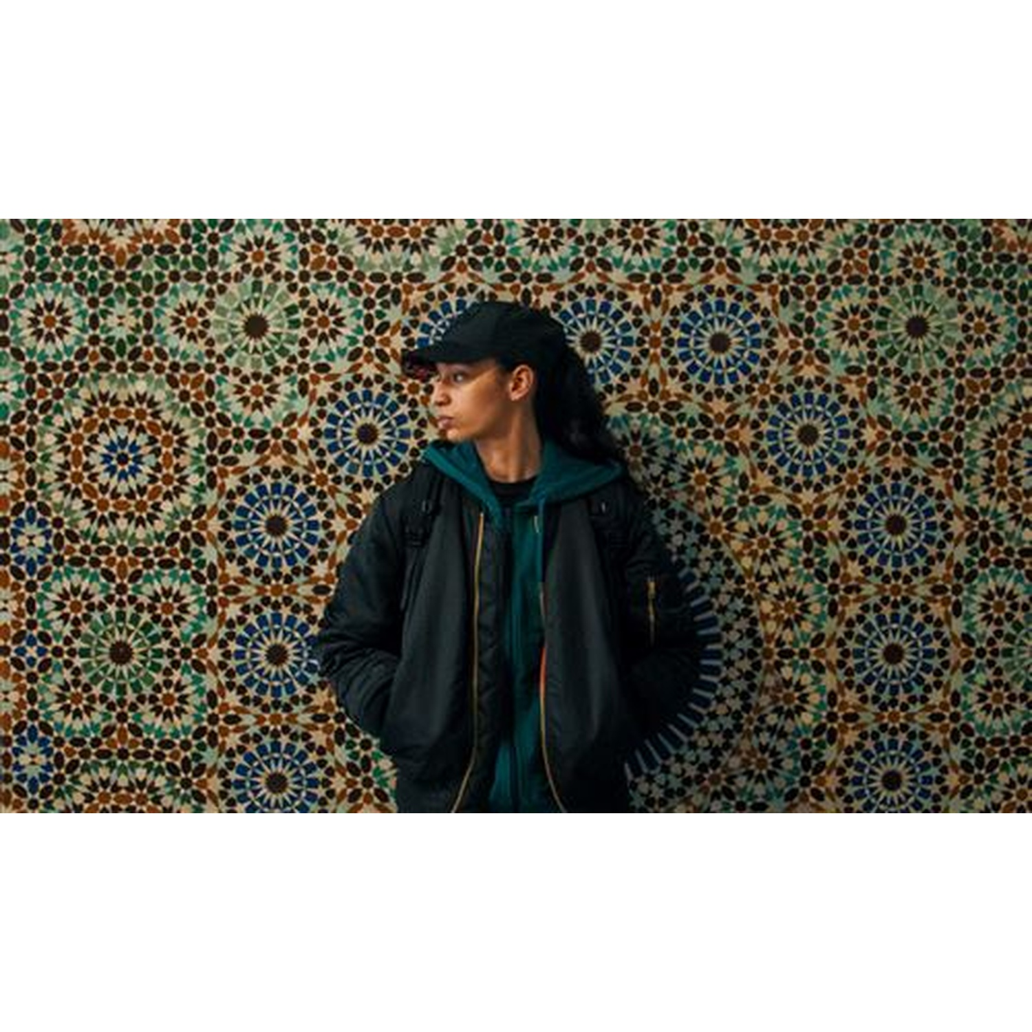 Une personne en sweat à capuche et casquette se tient devant un mur de mosaïque coloré.