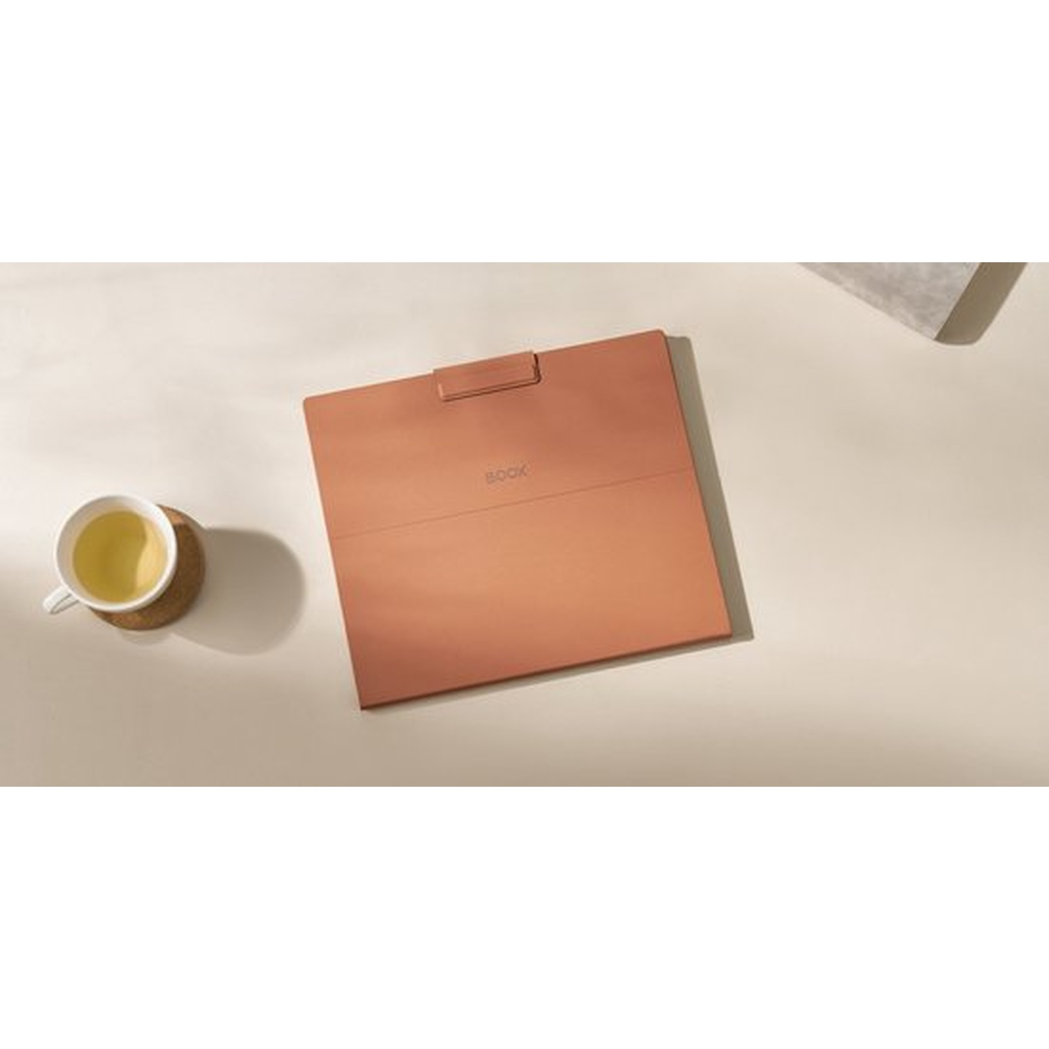 Een koperkleurige e-reader naast een kop thee op een lichte tafel.