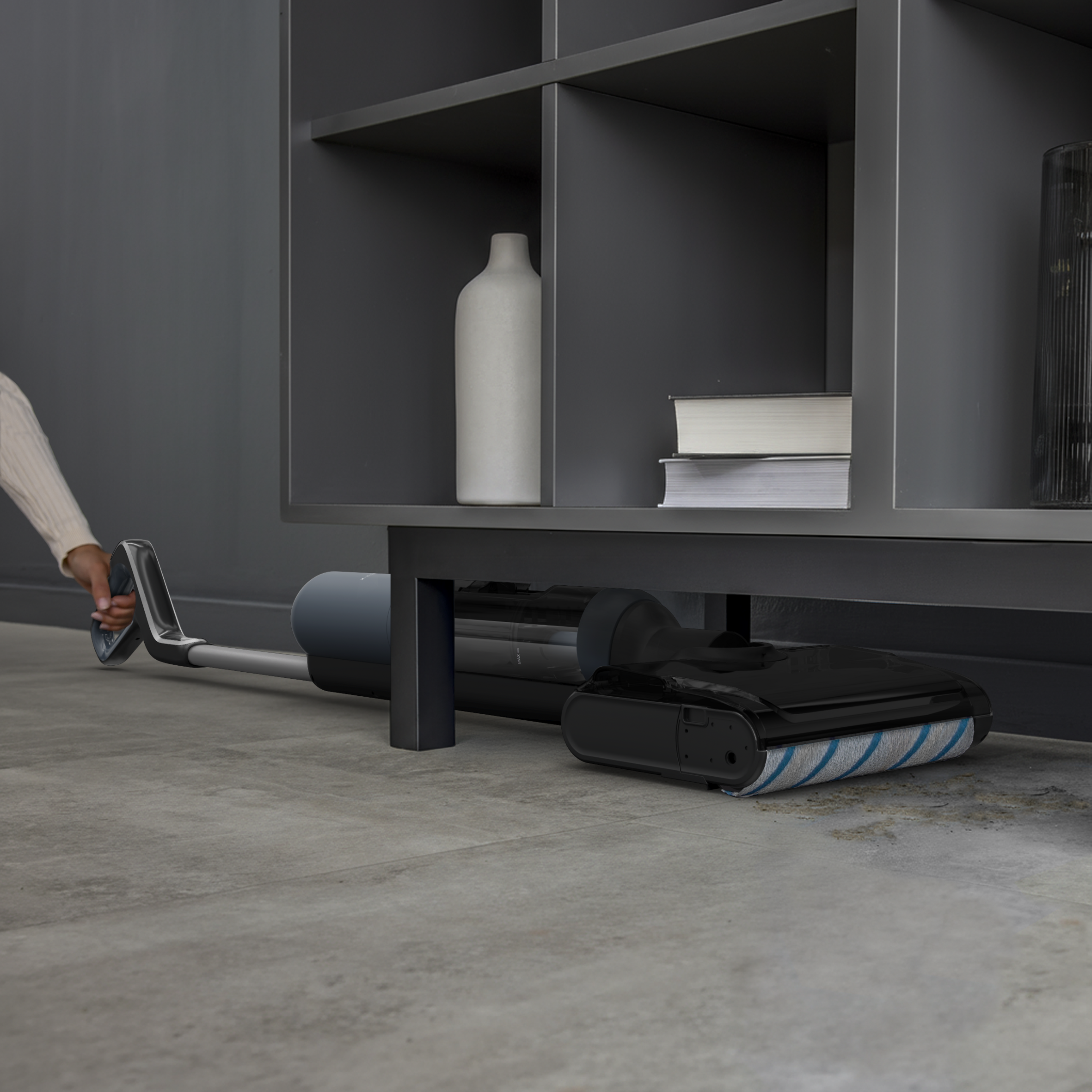 Une personne utilise un aspirateur sans fil sous une étagère. Fond sombre.