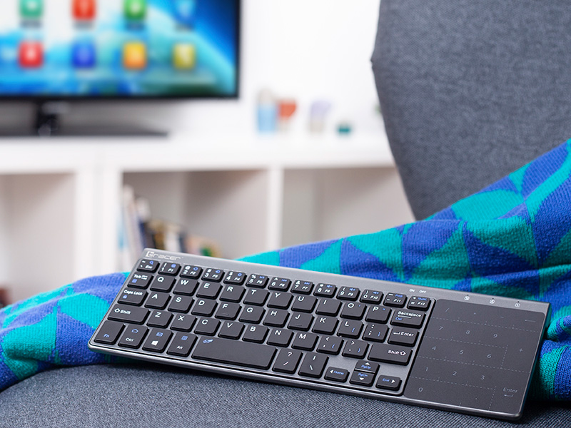 Klawiatura bezprzewodowa z touchpadem leży na niebiesko-zielonym kocu. W tle telewizor.