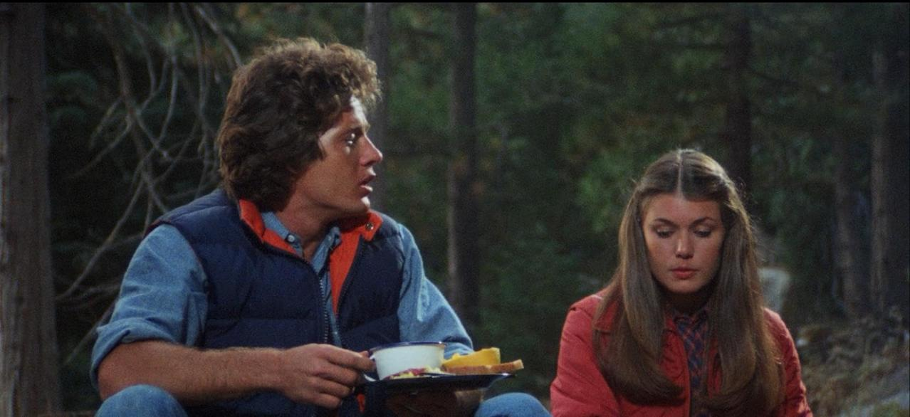 Ein Mann mit Locken und eine Frau sitzen im Wald. Er hält eine Tasse.