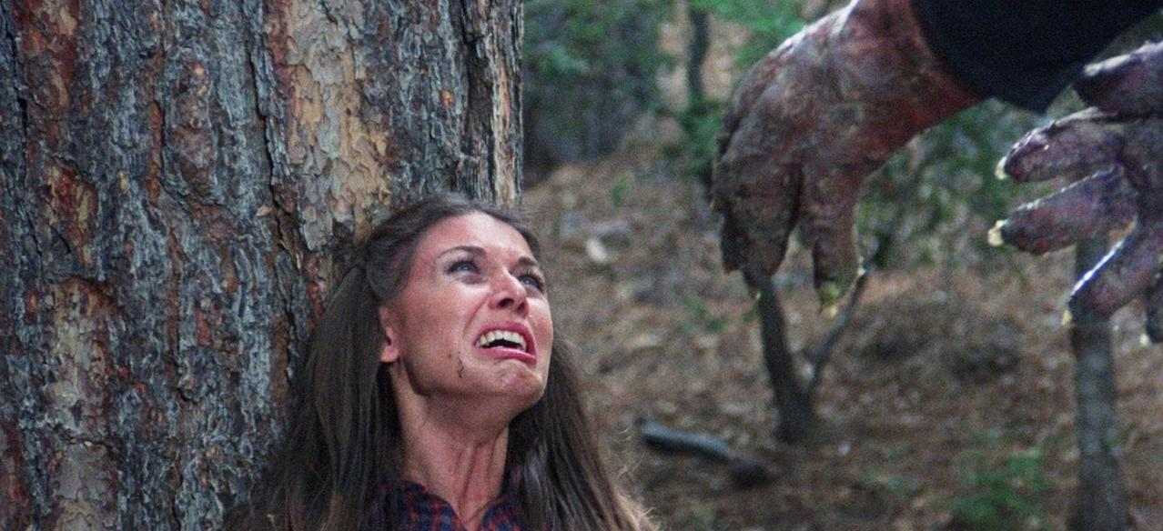 Eine schreiende Frau lehnt an einem Baum, während eine monströse Hand sich nähert.