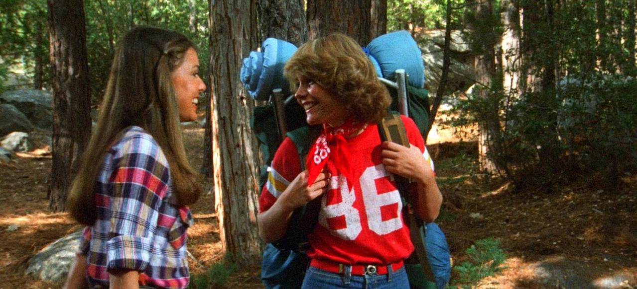 Zwei junge Frauen im Wald, eine mit kariertem Hemd, die andere mit rotem T-Shirt.