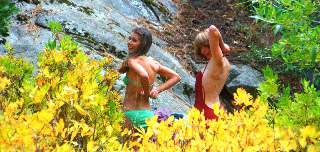 Zwei Frauen im Bikini zwischen gelben und grünen Blättern vor einem Felsen.