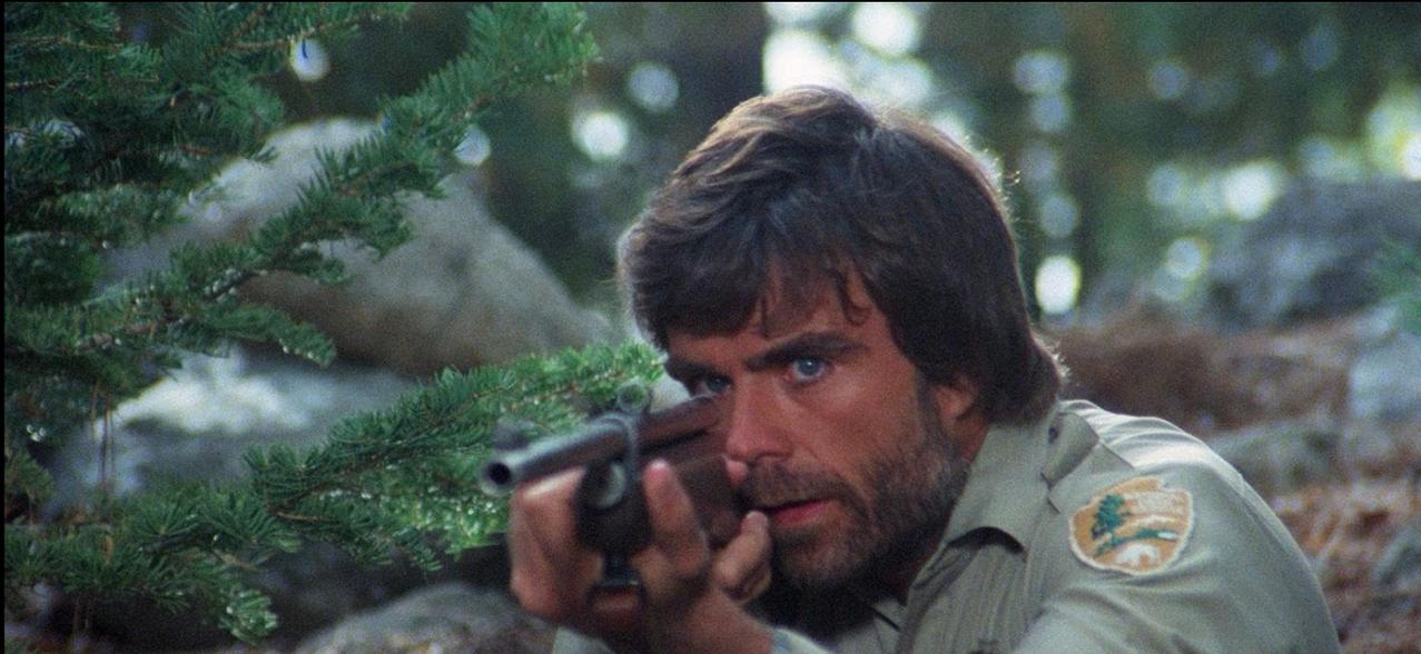 Ein Mann mit braunen Haaren und Bart zielt mit einem Gewehr durch Nadelbäume.