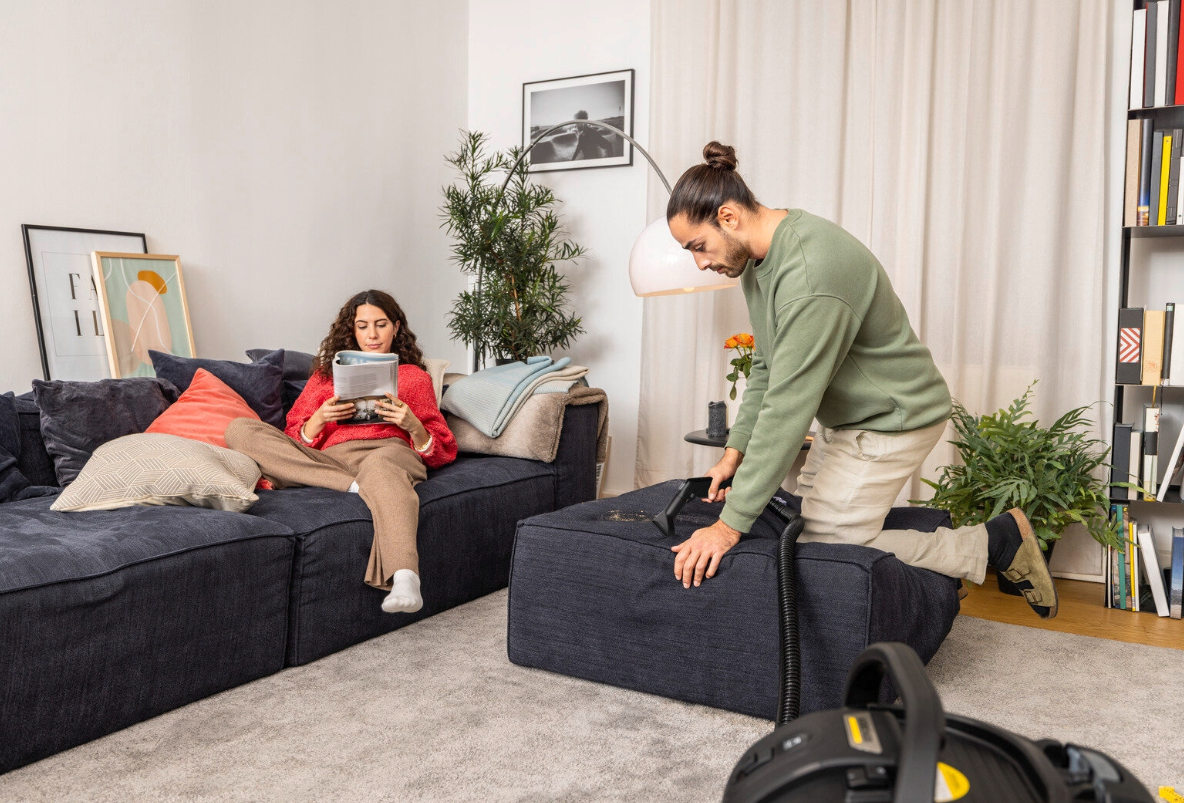 Mężczyzna odkurza sofę, kobieta czyta książkę.