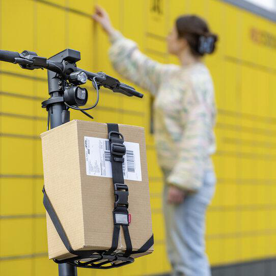 Ein Paket hängt an einem E-Scooter vor gelben Paketfächern, eine Frau greift danach.