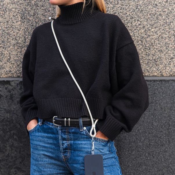 Eine Person mit schwarzem Rollkragenpullover, blauer Jeans und Handy am Trageband.