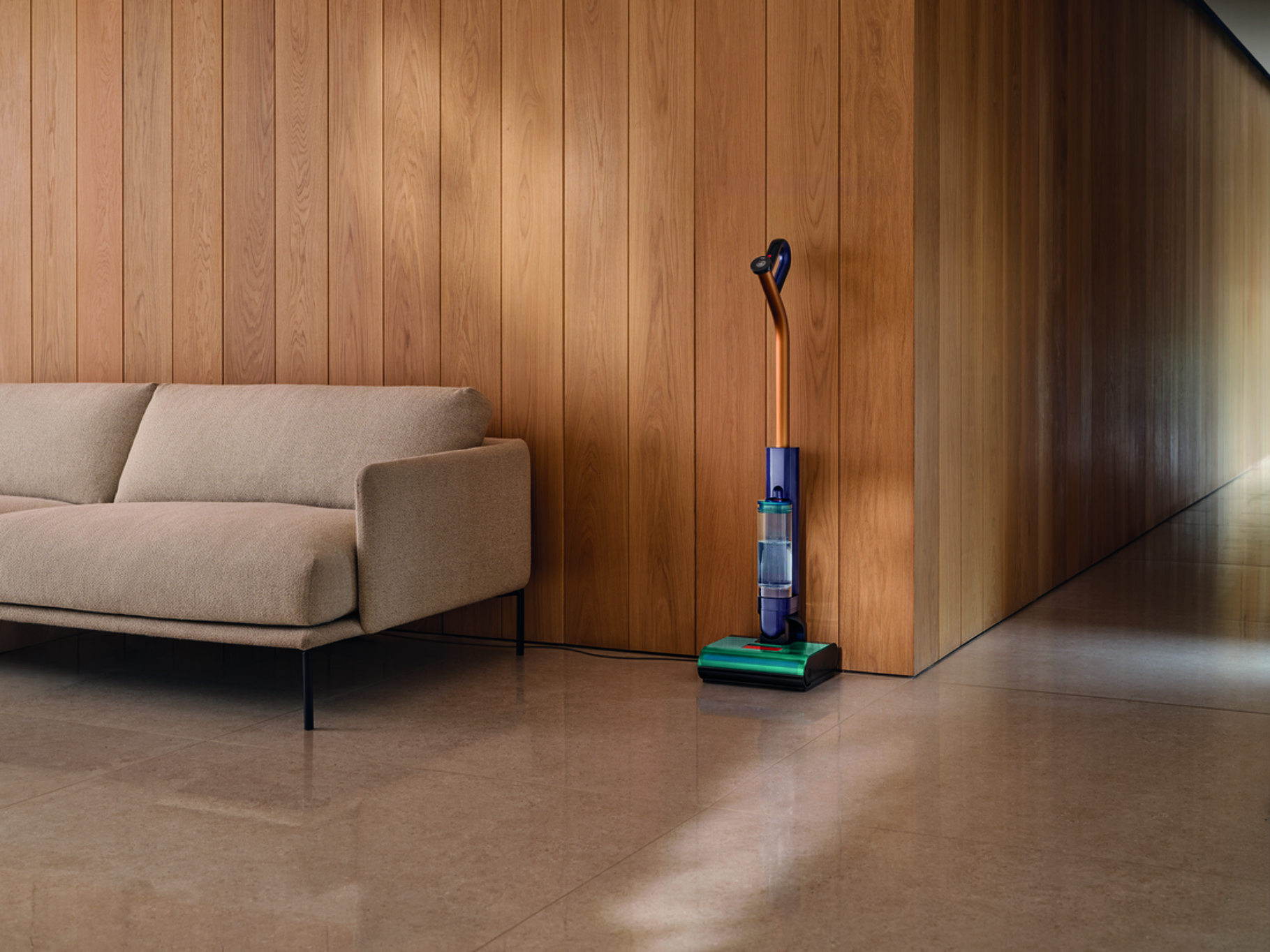 Beżowa sofa obok drewnianej ściany. Odkurzacz stoi na podłodze.
