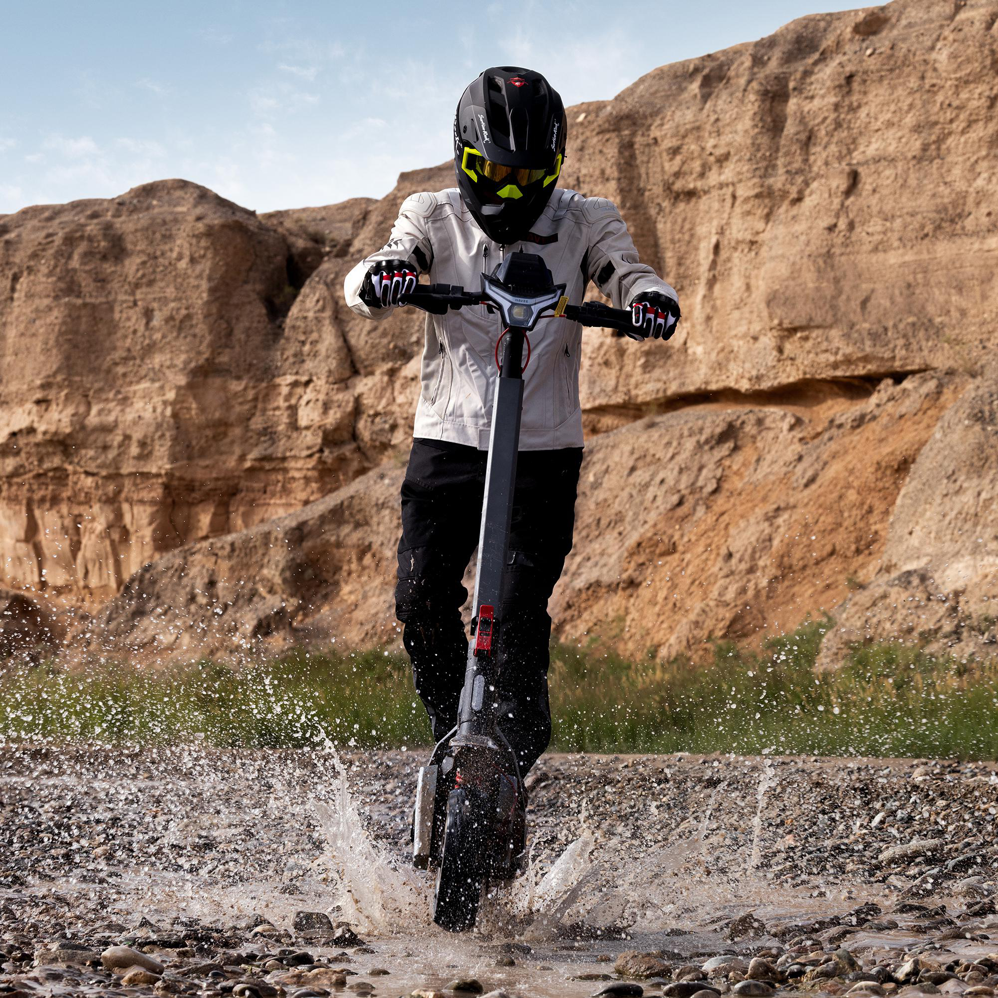 Fahrer mit Helm fährt E-Scooter durch eine Wasserlache vor Felsen.