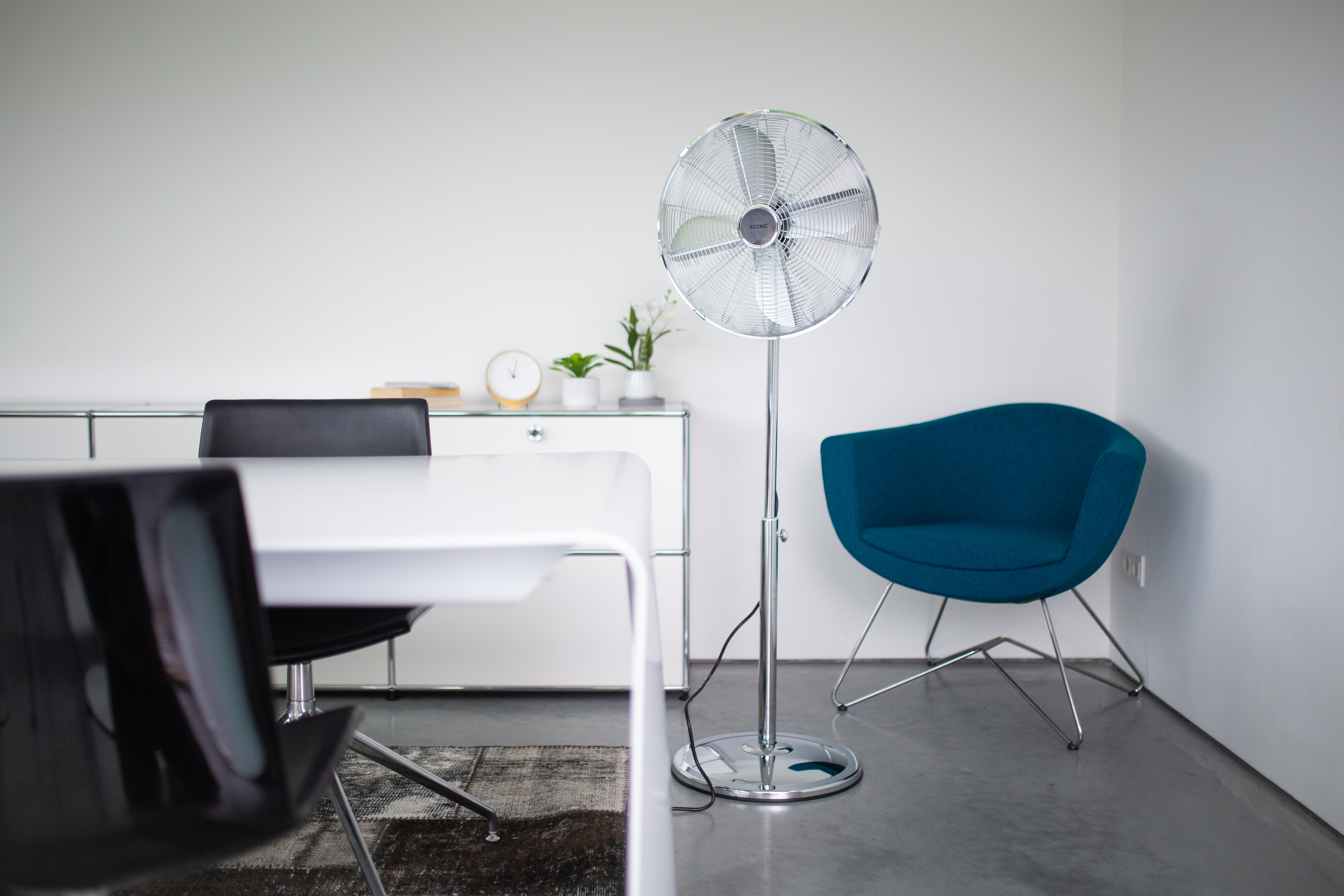 Pomieszczenie biurowe z białym stołem, czarnymi krzesłami, wentylatorem i niebieskim fotelem.