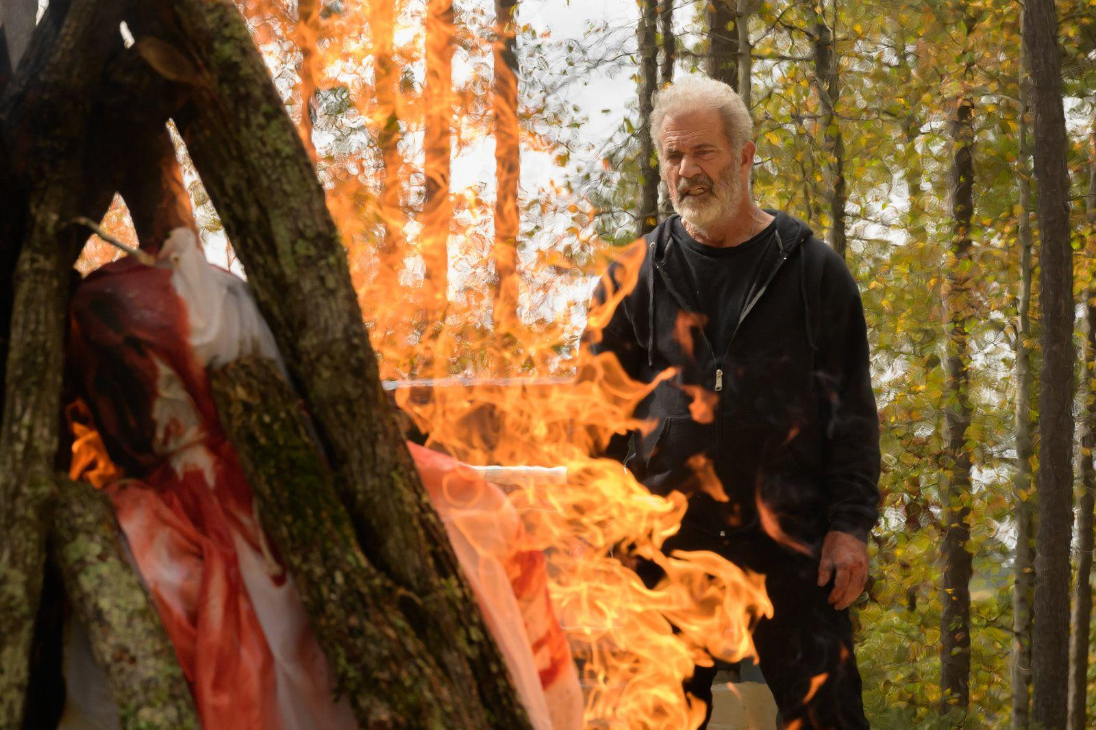 Mann in schwarzer Kleidung beobachtet Feuer in einem Wald mit Bäumen und Herbstlaub.