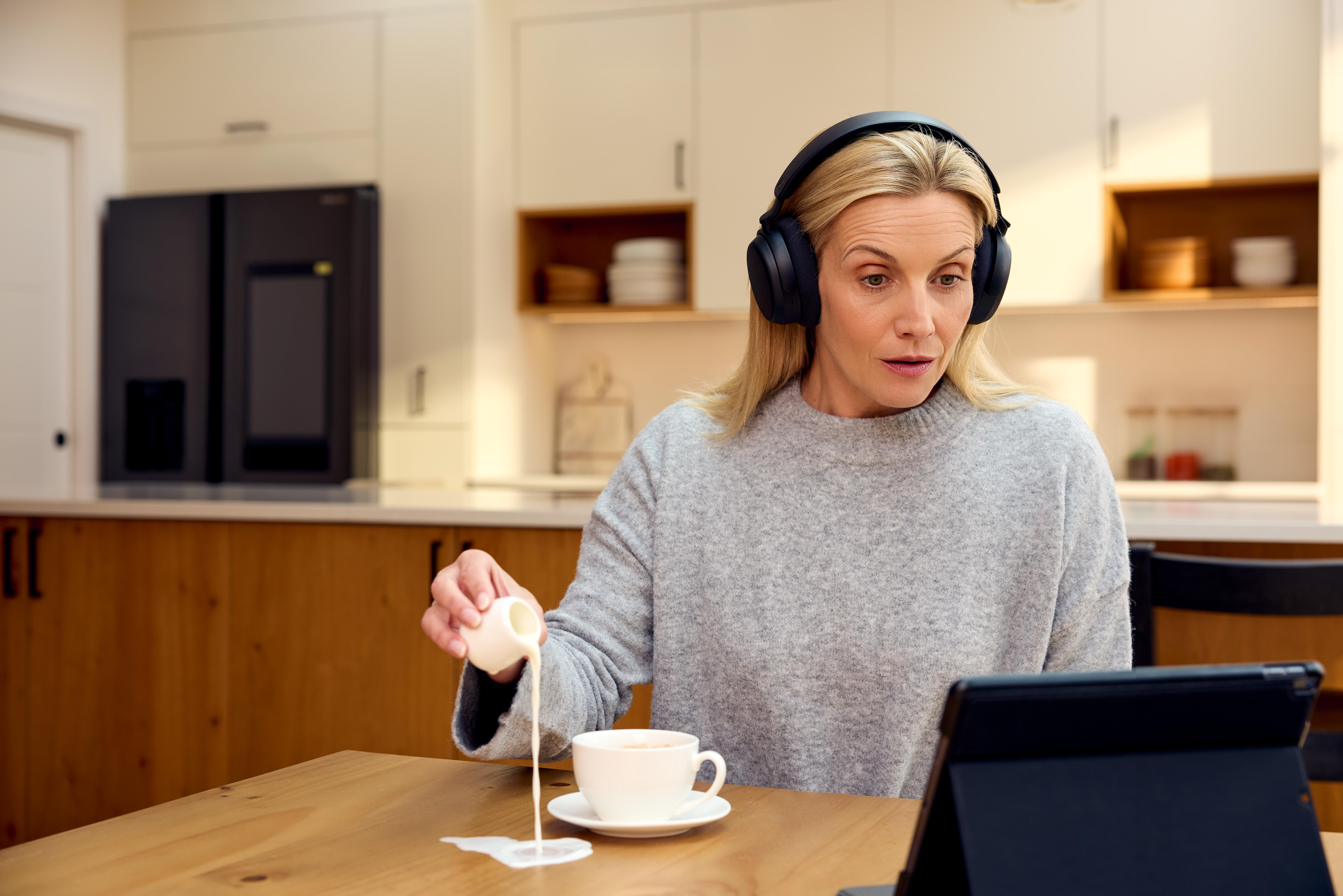 Eine Frau mit Kopfhörern gießt Milch in eine Tasse Kaffee, während sie auf ein Tablet schaut.
