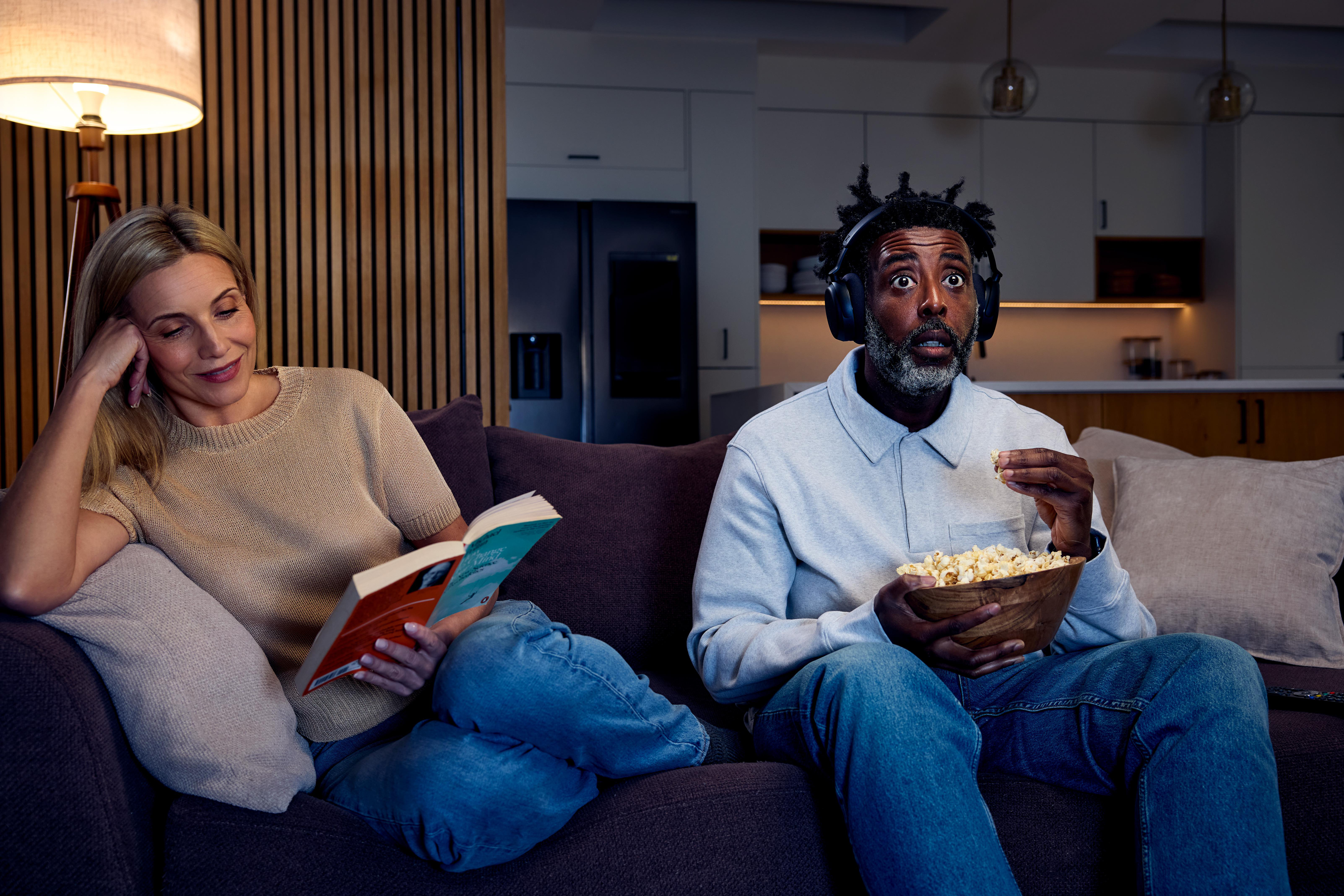 Mann mit Kopfhörern isst Popcorn, Frau liest Buch auf Couch.