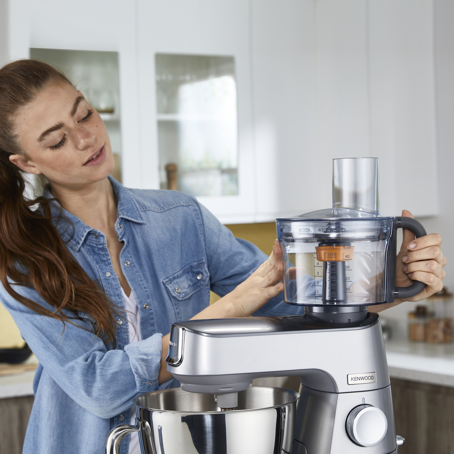 Una donna in camicia di jeans usa un robot da cucina con ciotola argentata e contenitore trasparente.