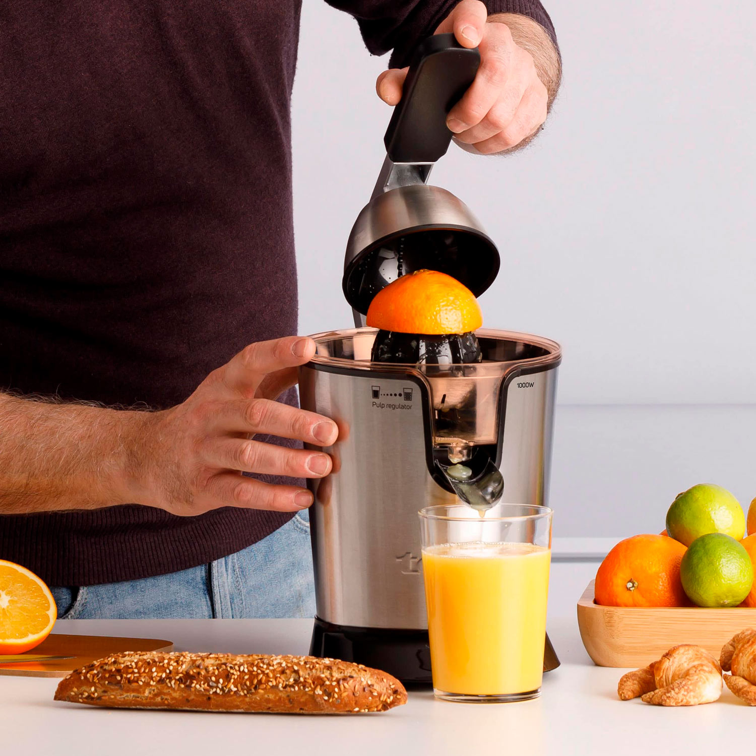 Hombre exprimiendo una naranja. El zumo fluye hacia un vaso. Frutas y pan cerca.