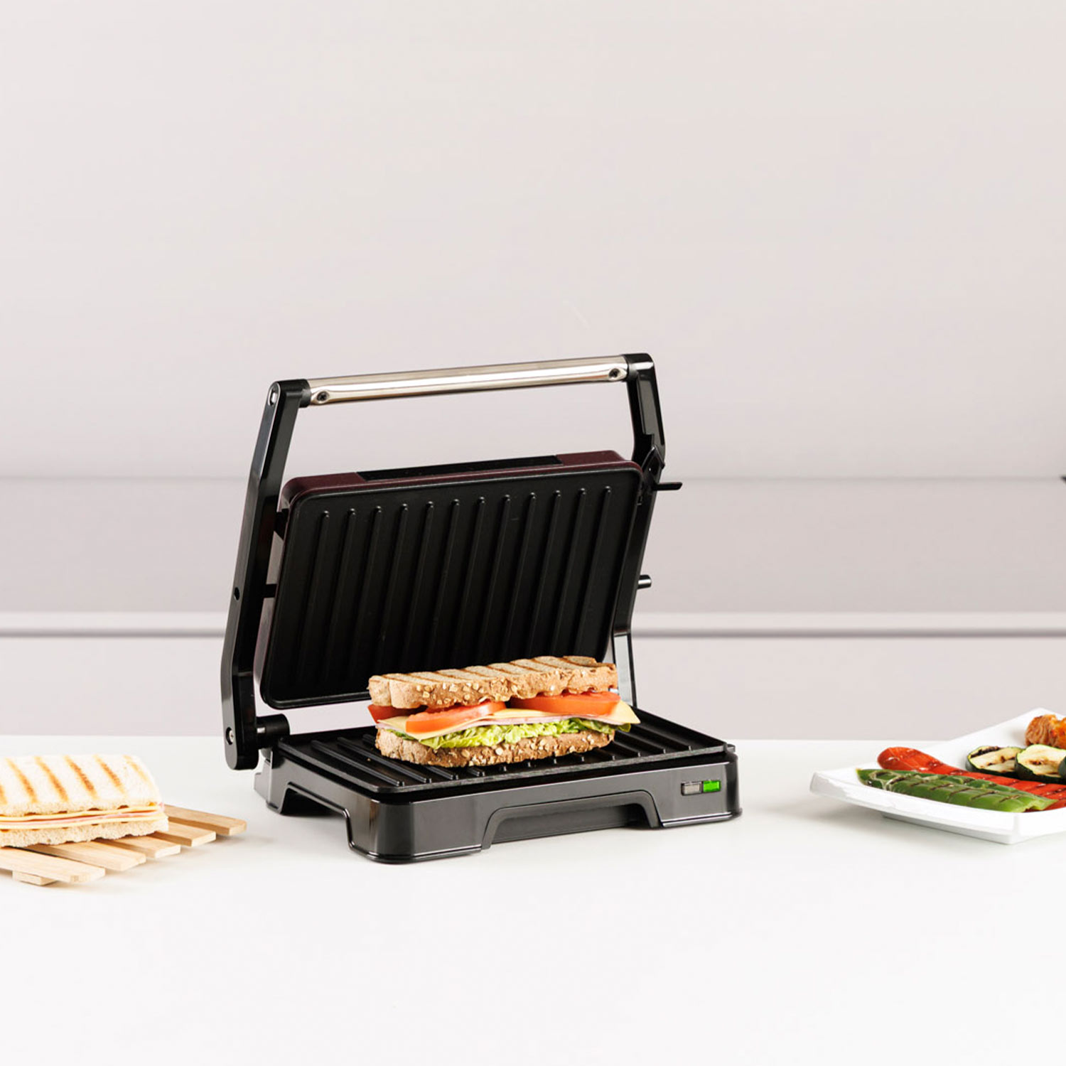 Una sandwichera abierta con un sándwich, al lado otro sándwich y verduras a la parrilla.