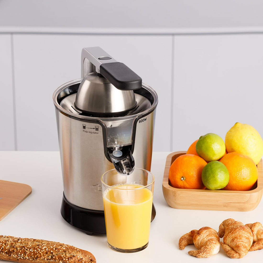 Un exprimidor con vaso de zumo, frutas y bollería en una encimera.