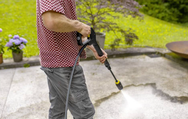 Mężczyzna myje kamienny taras myjką ciśnieniową. Zieleń i rośliny w tle.