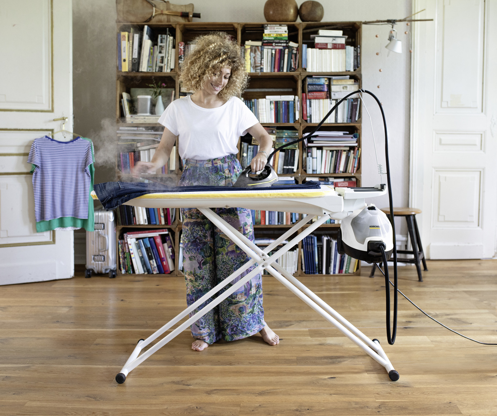 Kobieta prasuje spodnie na desce, unosi się para. Regał z książkami w tle.