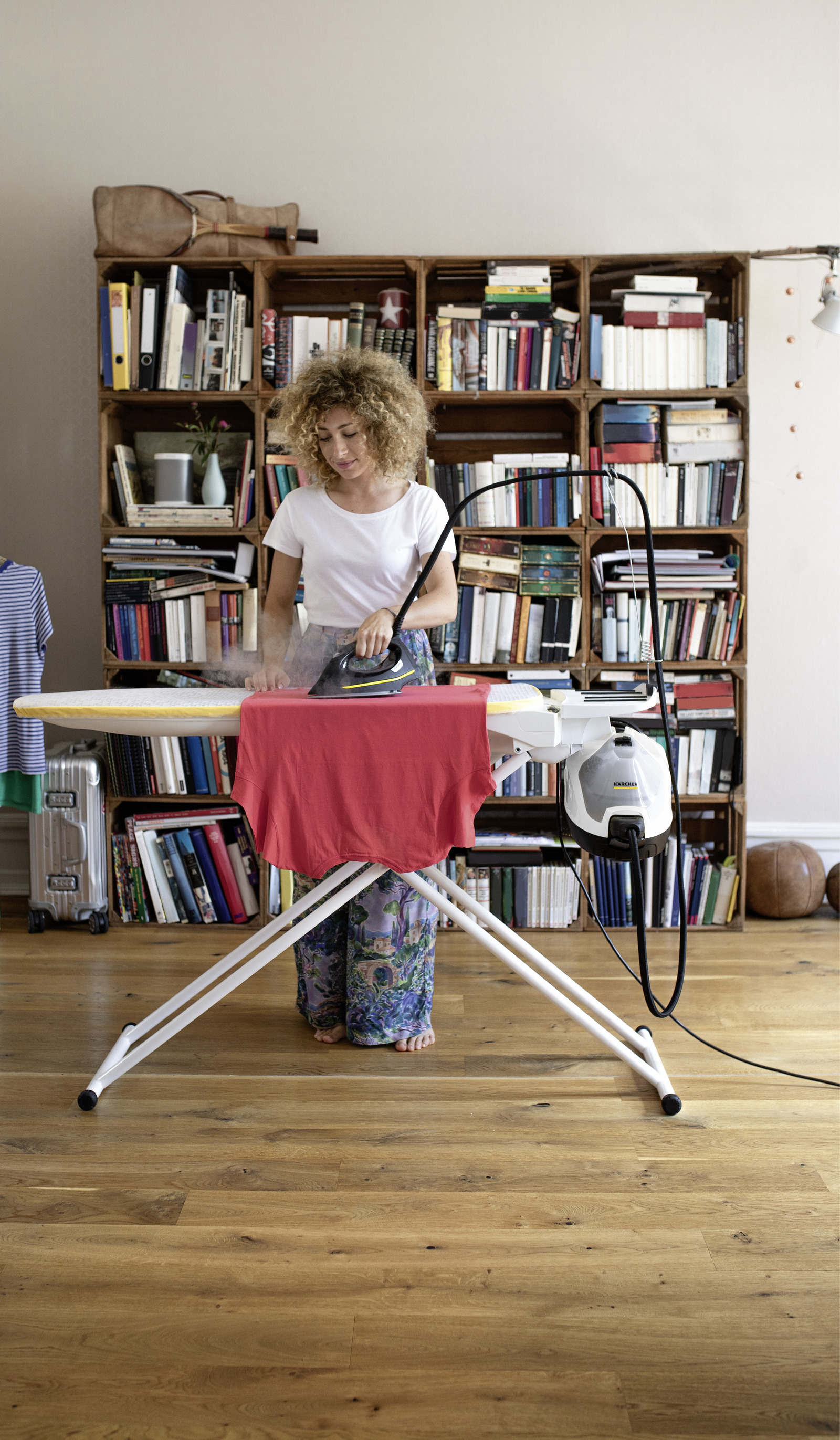 Kobieta prasuje czerwoną koszulę na desce do prasowania przed regałem z książkami.