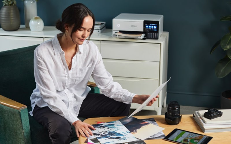 Kobieta w białej koszuli przegląda papiery. Drukarka stoi na komodzie.