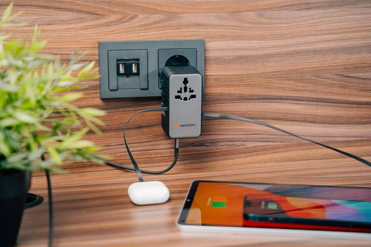 Egy utazási adapter van a fali aljzatba dugva, töltve egy táblagépet és fehér fülhallgatót.