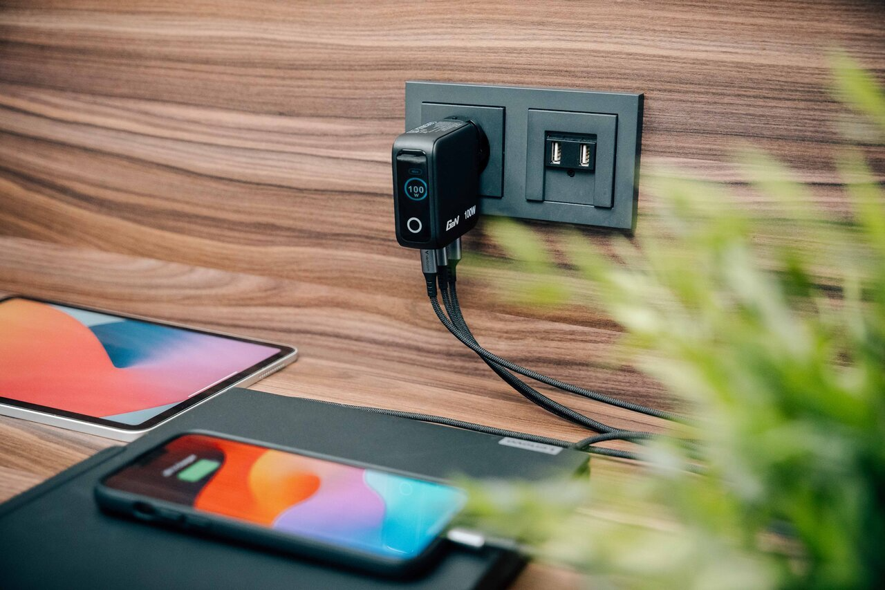 Egy fekete 100W-os töltő csatlakoztatva fali aljzathoz. Közelben tablet és telefon.