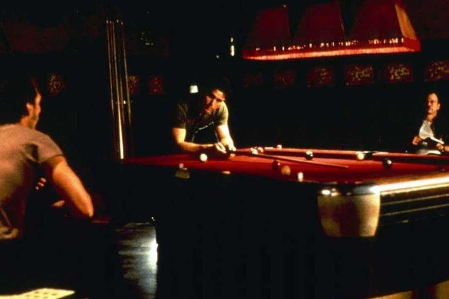 Drei Männer spielen Billard in einem schwach beleuchteten Raum. Ein Mann zielt auf den roten Filztisch.