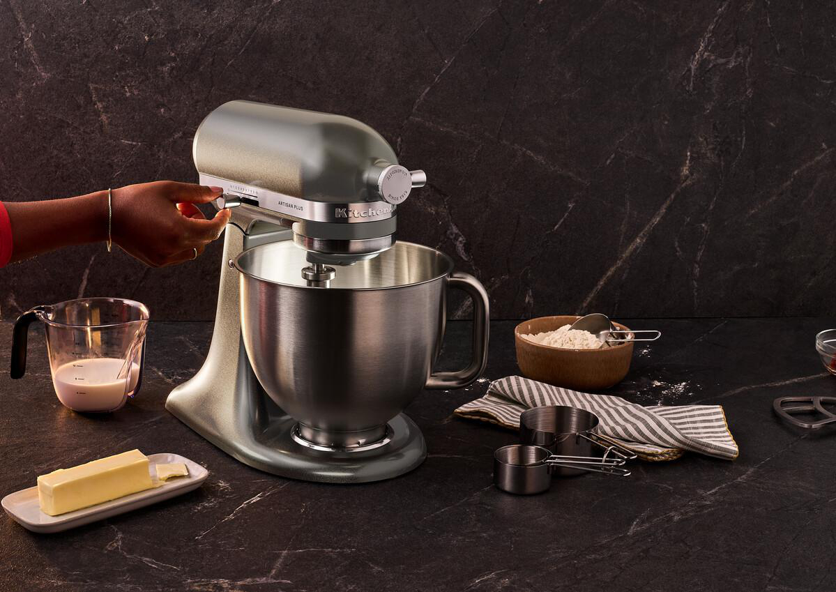 Eine Hand stellt eine Küchenmaschine mit Edelstahlschüssel ein. Butter, Milch, Mehl und Messbecher sind um sie herum.
