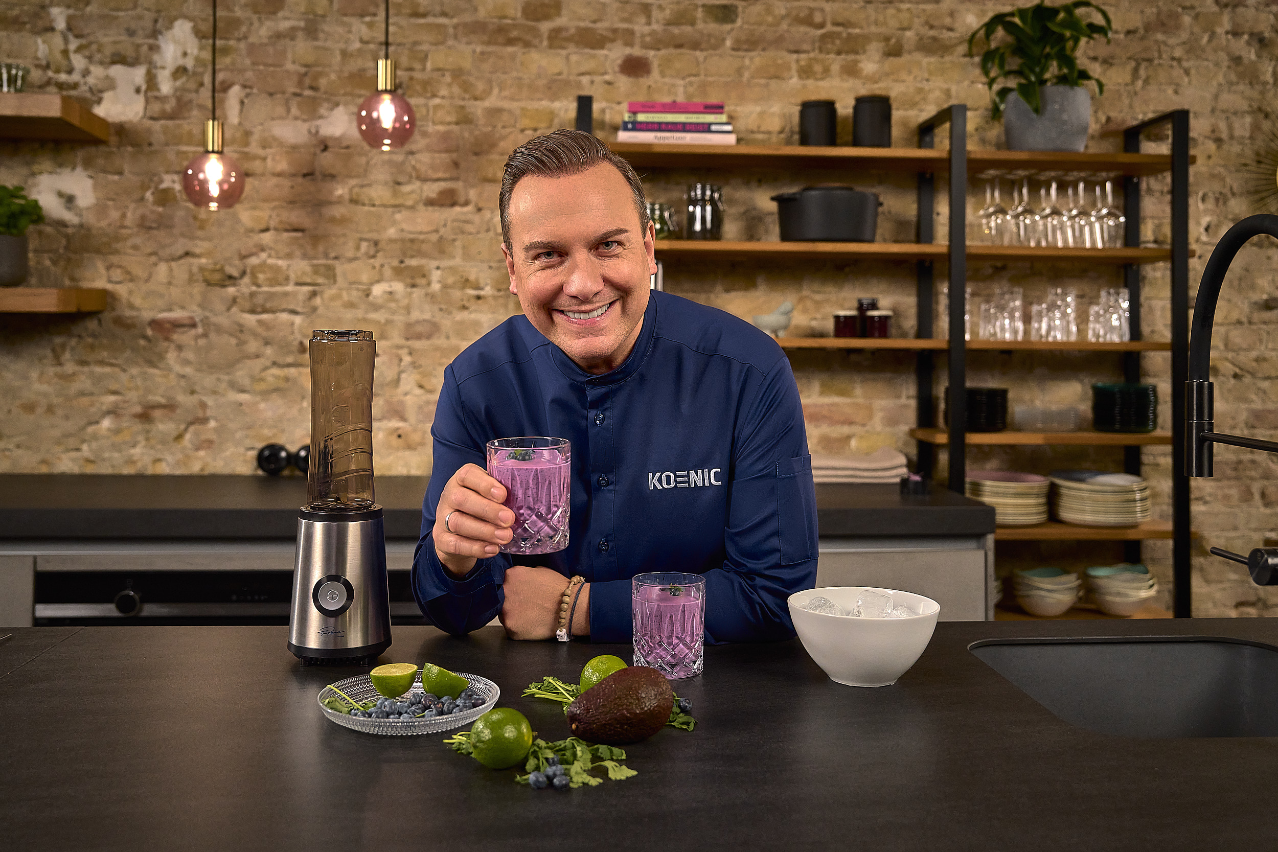 Mężczyzna w granatowej koszuli trzyma szklankę fioletowego smoothie. Obok stoi blender.