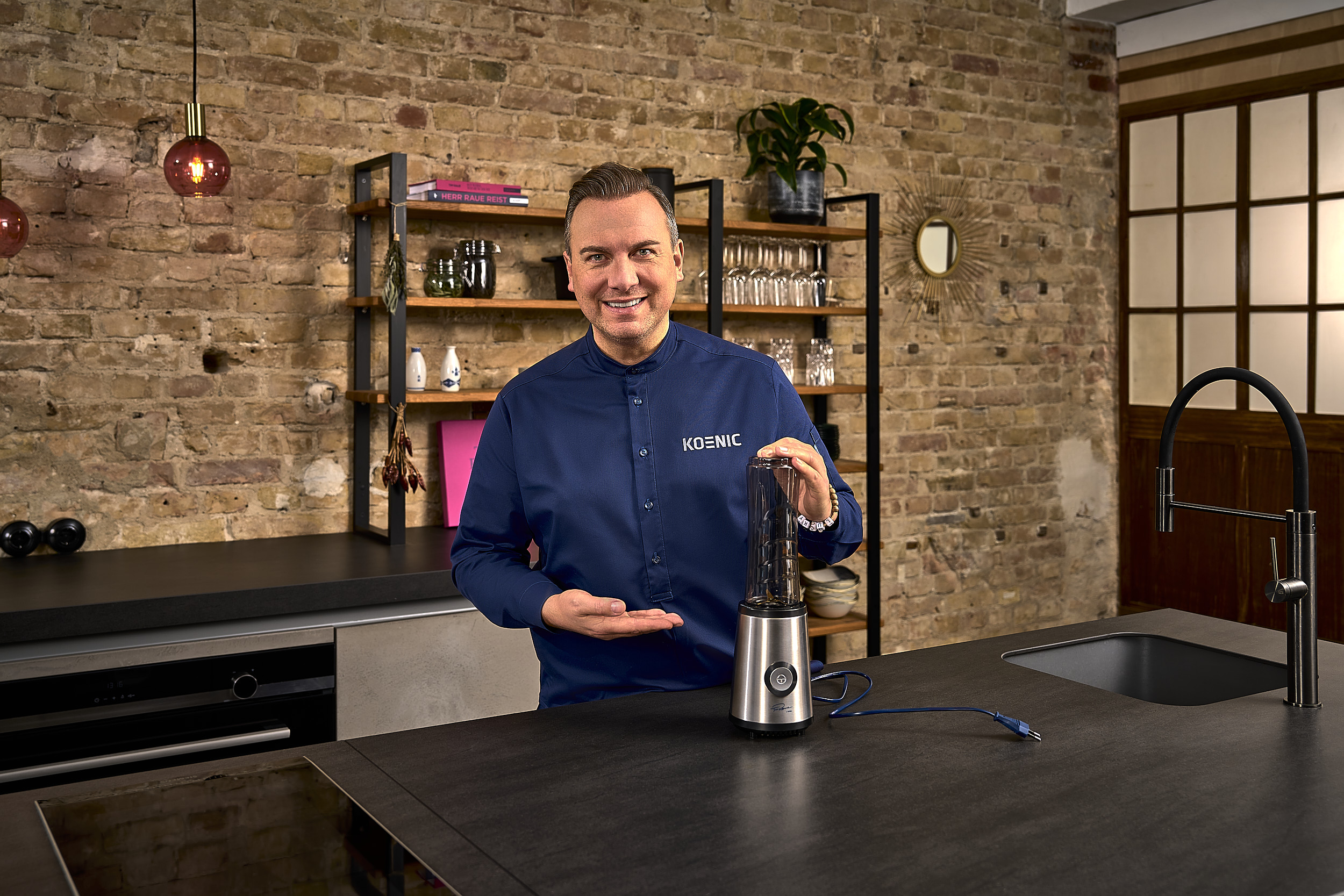 Mężczyzna w niebieskiej koszuli prezentuje blender w nowoczesnej kuchni z ceglanym murem.