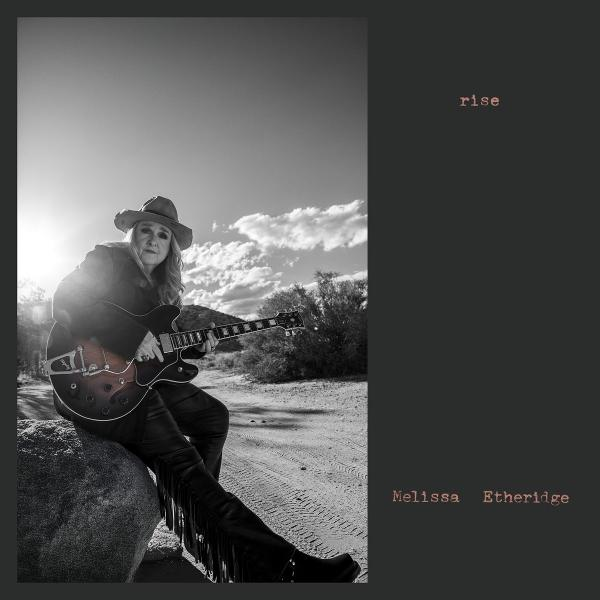 Eine Frau mit Hut spielt Gitarre im Freien, mit Felsen und Büschen im Hintergrund.