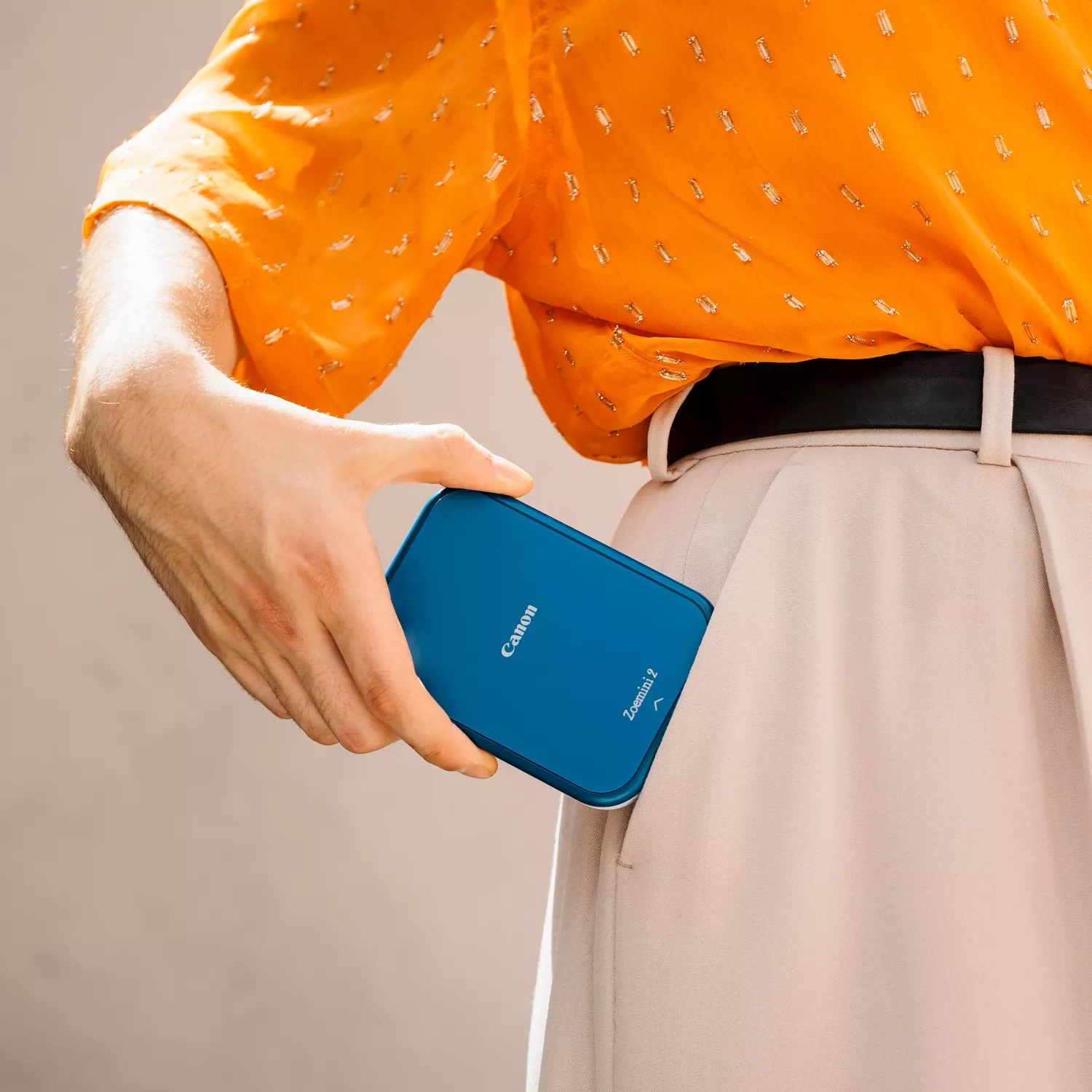 Ręka wkłada niebieską drukarkę Canon Zoemini 2 do jasnobeżowej kieszeni.