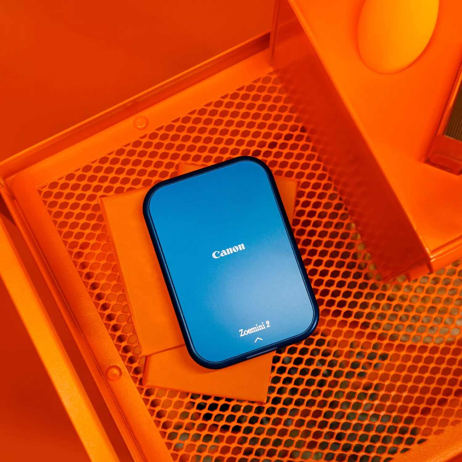 Niebieska drukarka Canon Zoemini 2 leży na pomarańczowej półce z siatki.