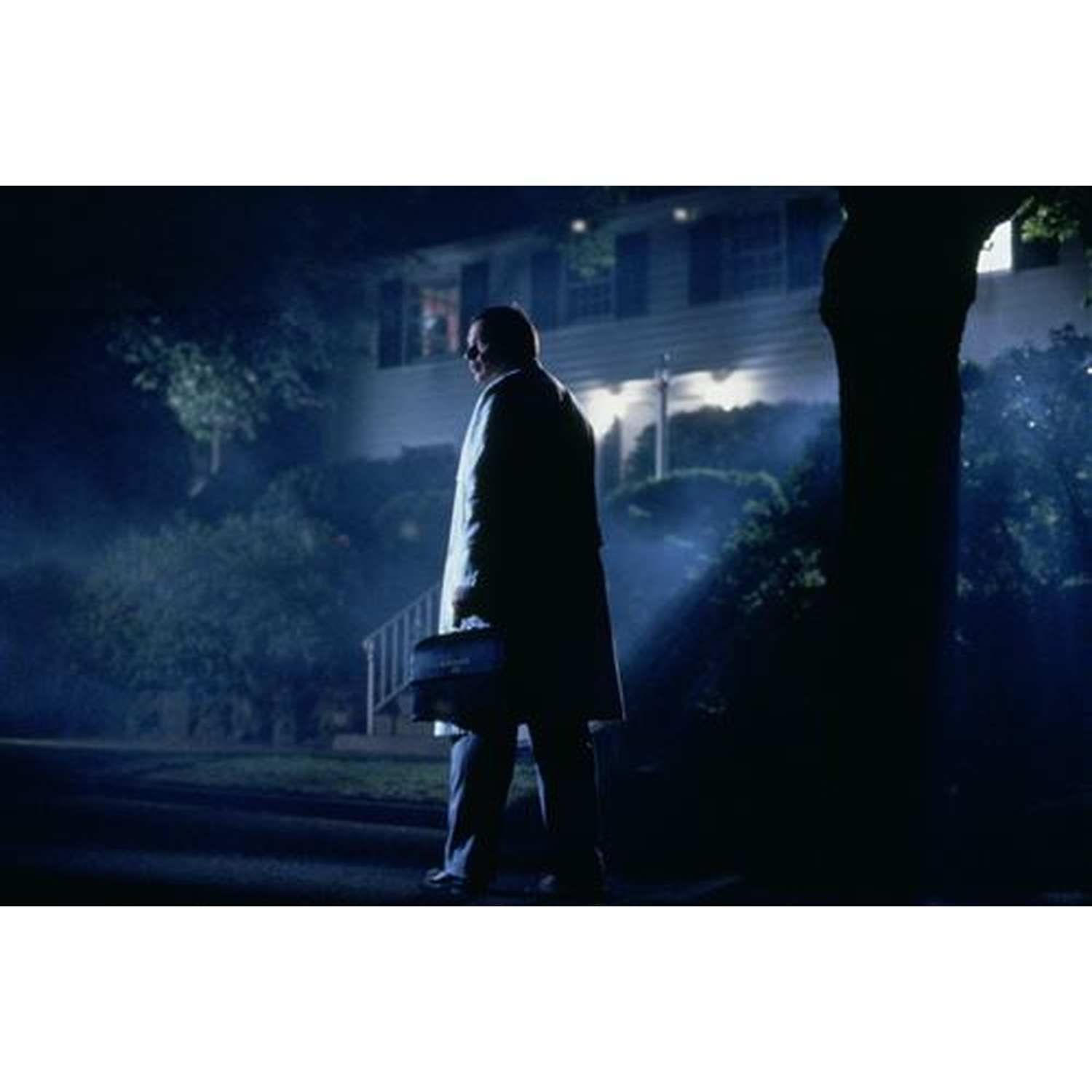 Homme en manteau sombre porte une mallette devant une maison illuminée la nuit.