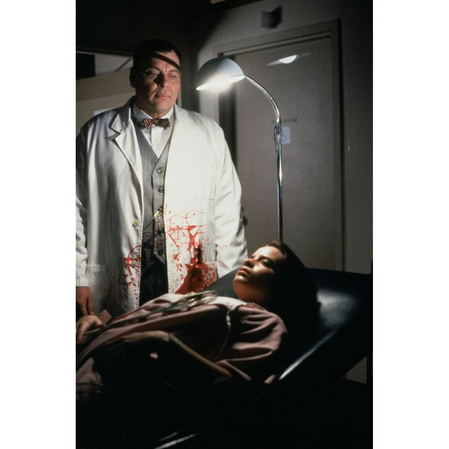 Un homme en blouse blanche avec des taches rouges se tient au-dessus d'une femme allongée sur un lit.
