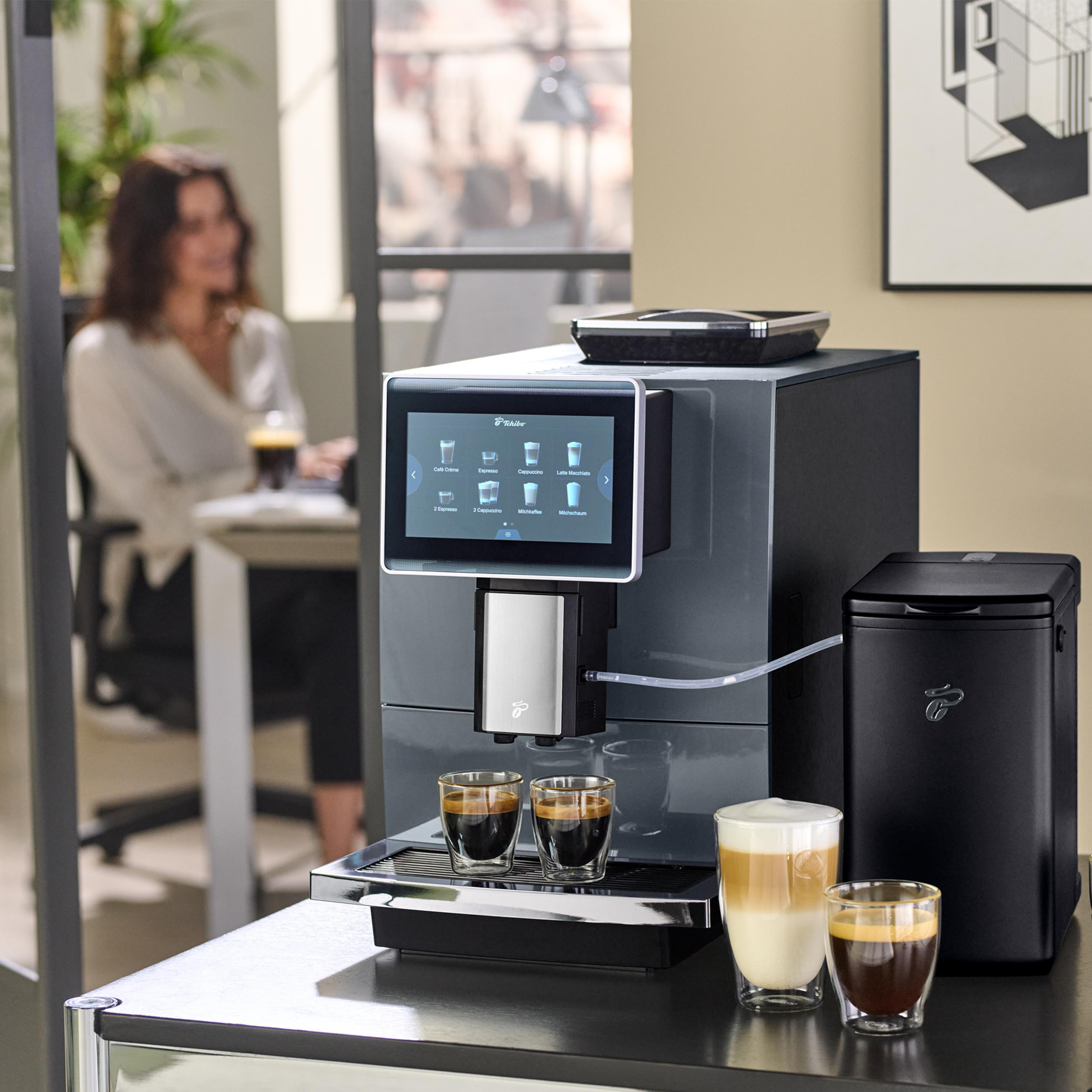 Kaffeemaschine mit zwei Espressos und einem Latte Macchiato. Frau im Hintergrund.
