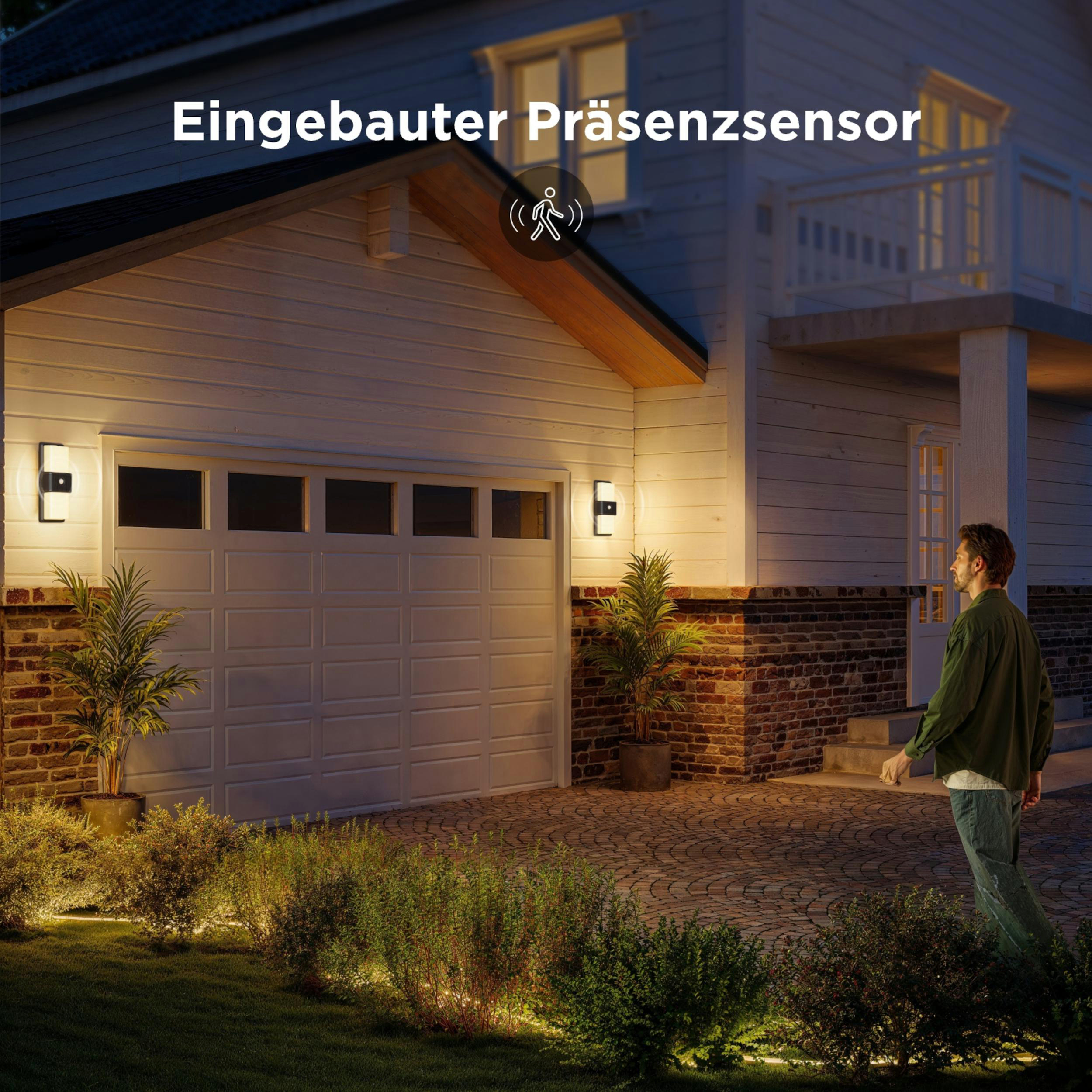 Ein Mann geht nachts auf eine beleuchtete Einfahrt zu einem Haus mit Garagentor.