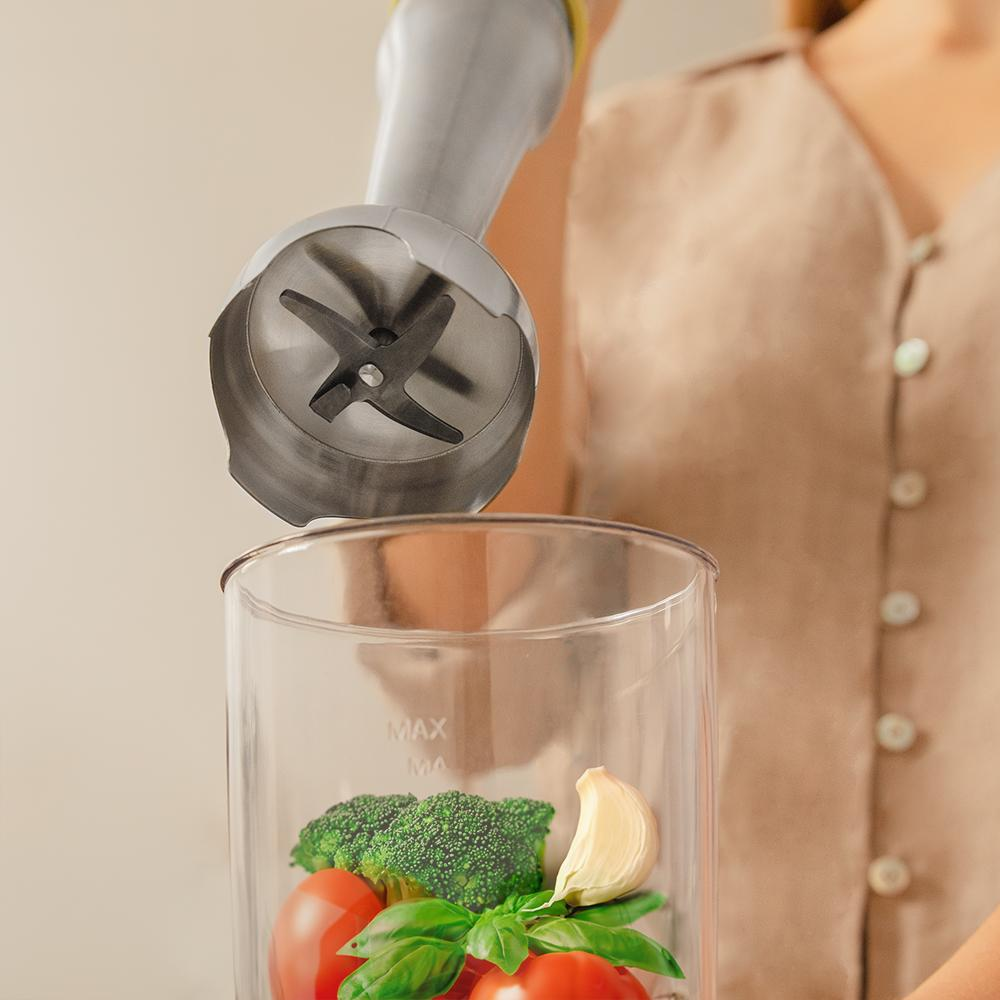 Batidora de mano a punto de mezclar brócoli, tomates, albahaca y ajo en un recipiente.
