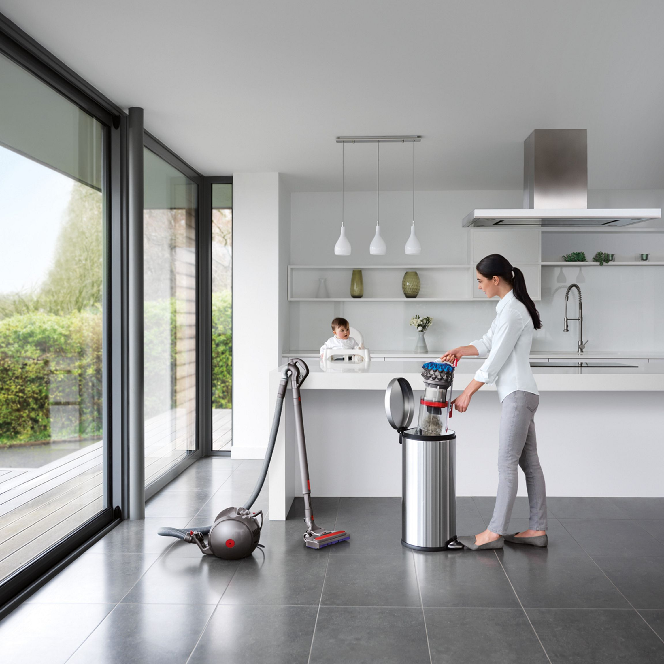 Une femme vide un aspirateur dans une poubelle dans une cuisine moderne avec un bébé dans une chaise haute à proximité et de grandes fenêtres.