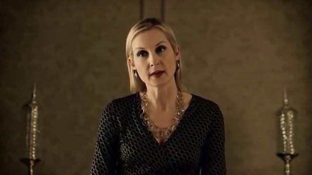 Eine blonde Frau in einem schwarzen Kleid mit Muster und Halskette steht vor einer Wand.