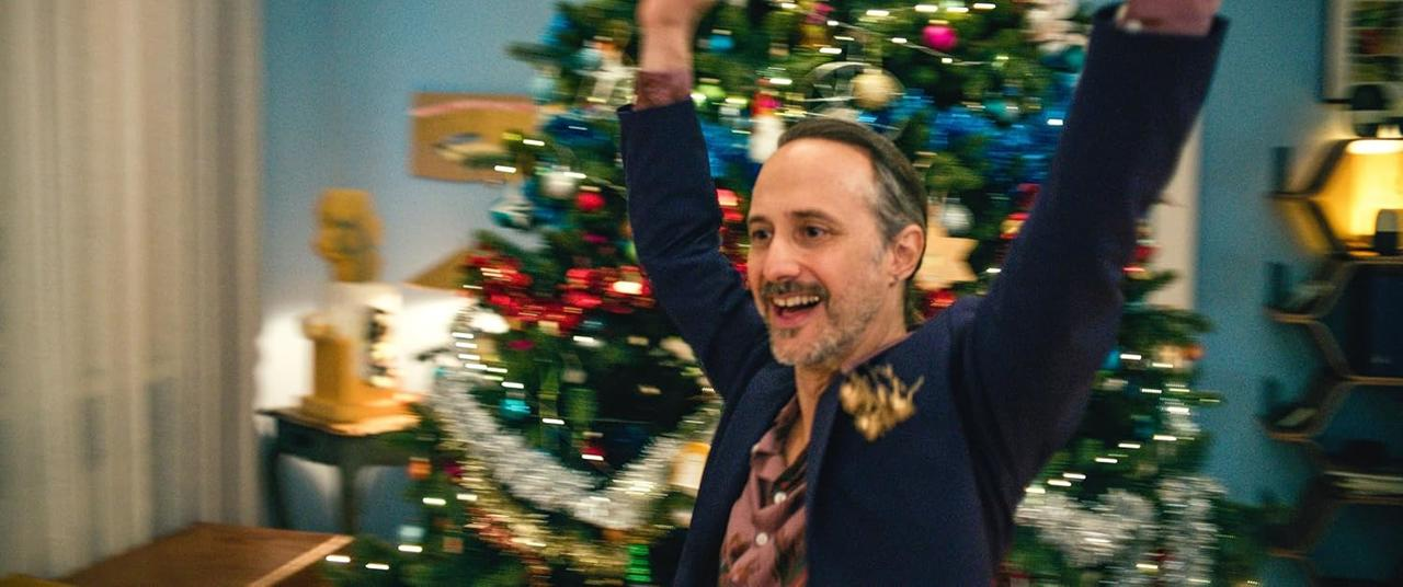 Ein Mann mit erhobenen Armen steht vor einem Weihnachtsbaum mit Lichtern.