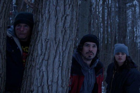 Drei Personen verstecken sich im winterlichen Wald hinter Bäumen.