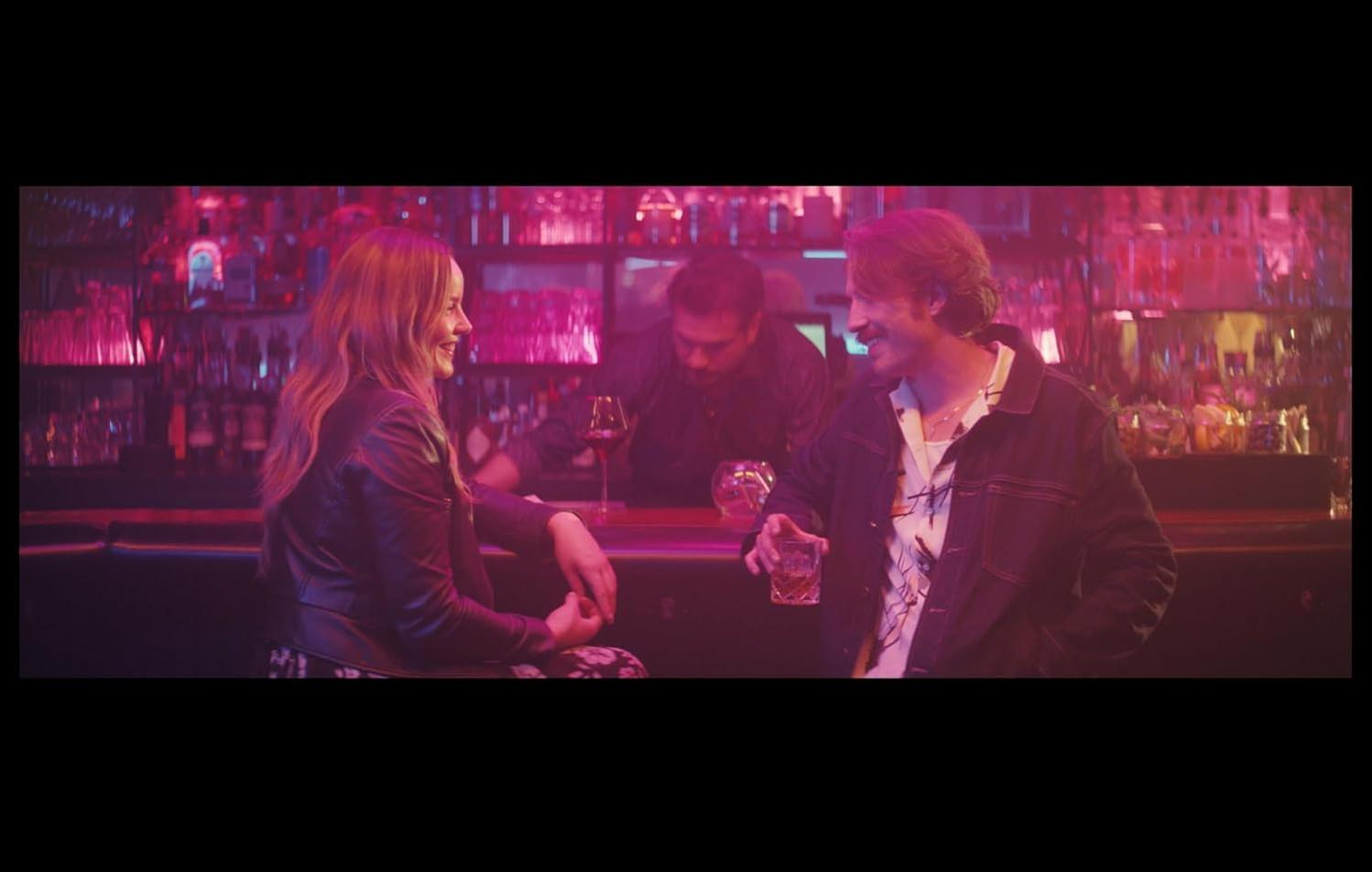 Ein Mann und eine Frau sitzen an einer Bar im Neonlicht. Ein Barkeeper ist im Hintergrund.
